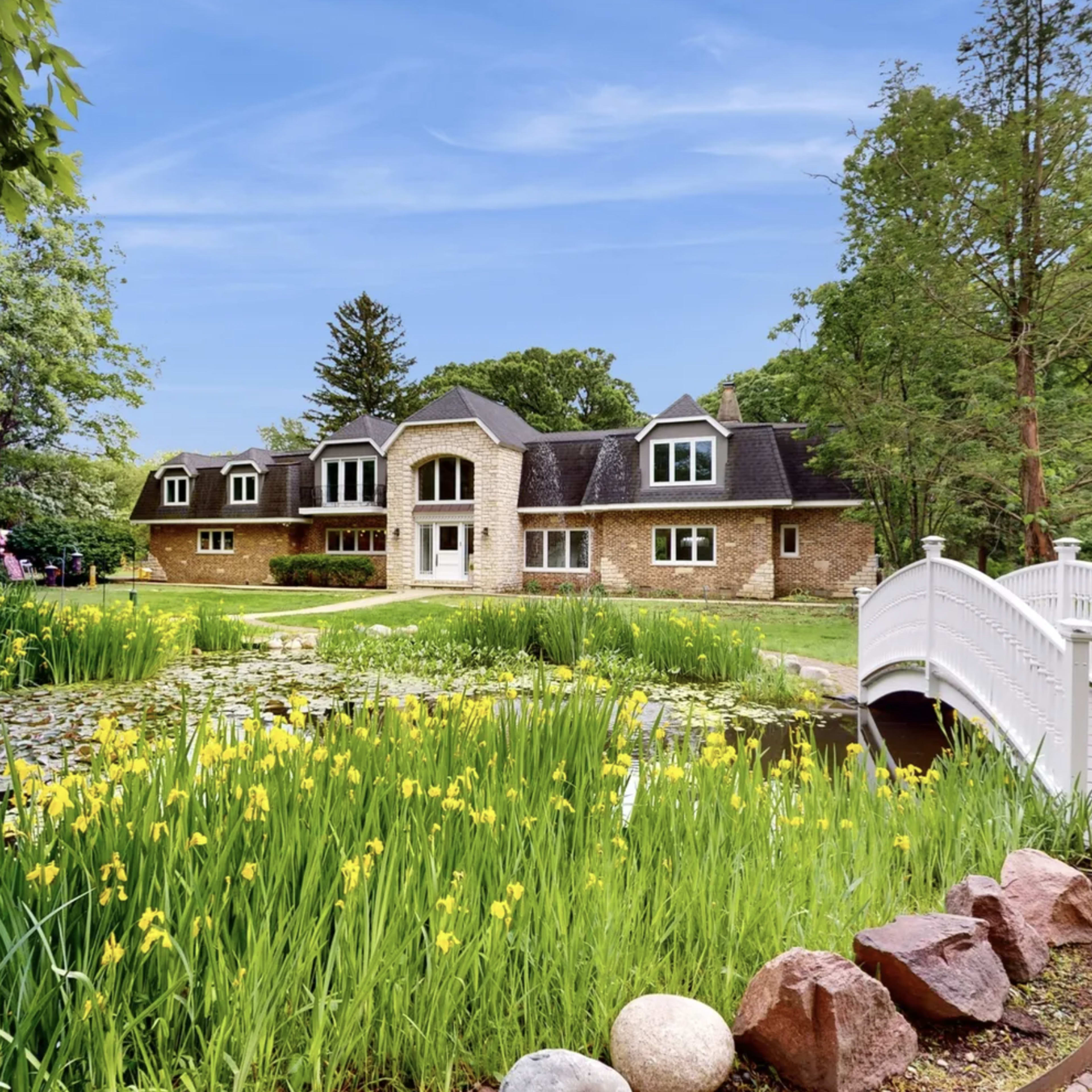 A stone house with multiple gabled roofs is situated near a pond surrounded by greenery and yellow flowers, with a white wooden bridge in the foreground.