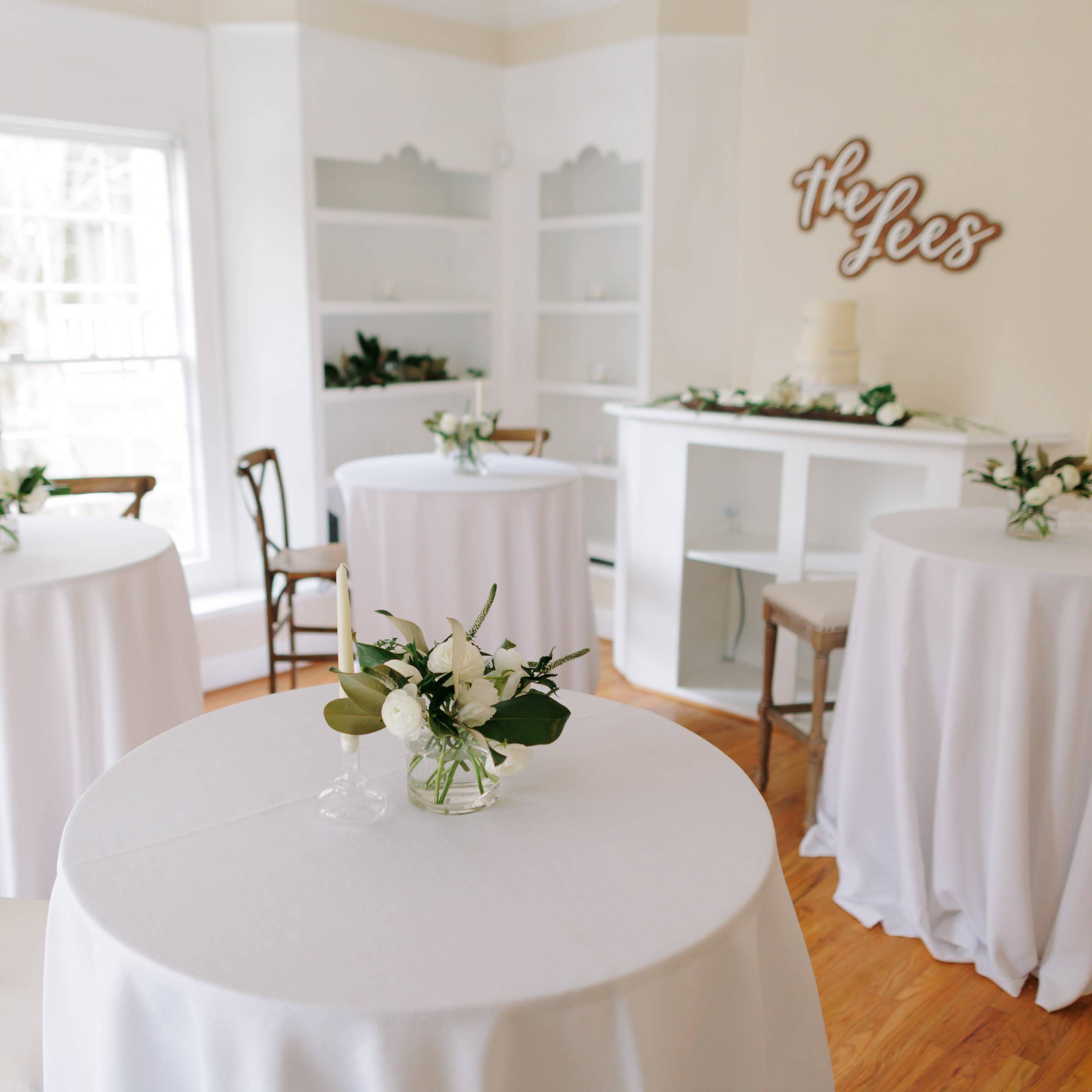 A light-filled room with several round tables covered in white tablecloths, each adorned with floral centerpieces.