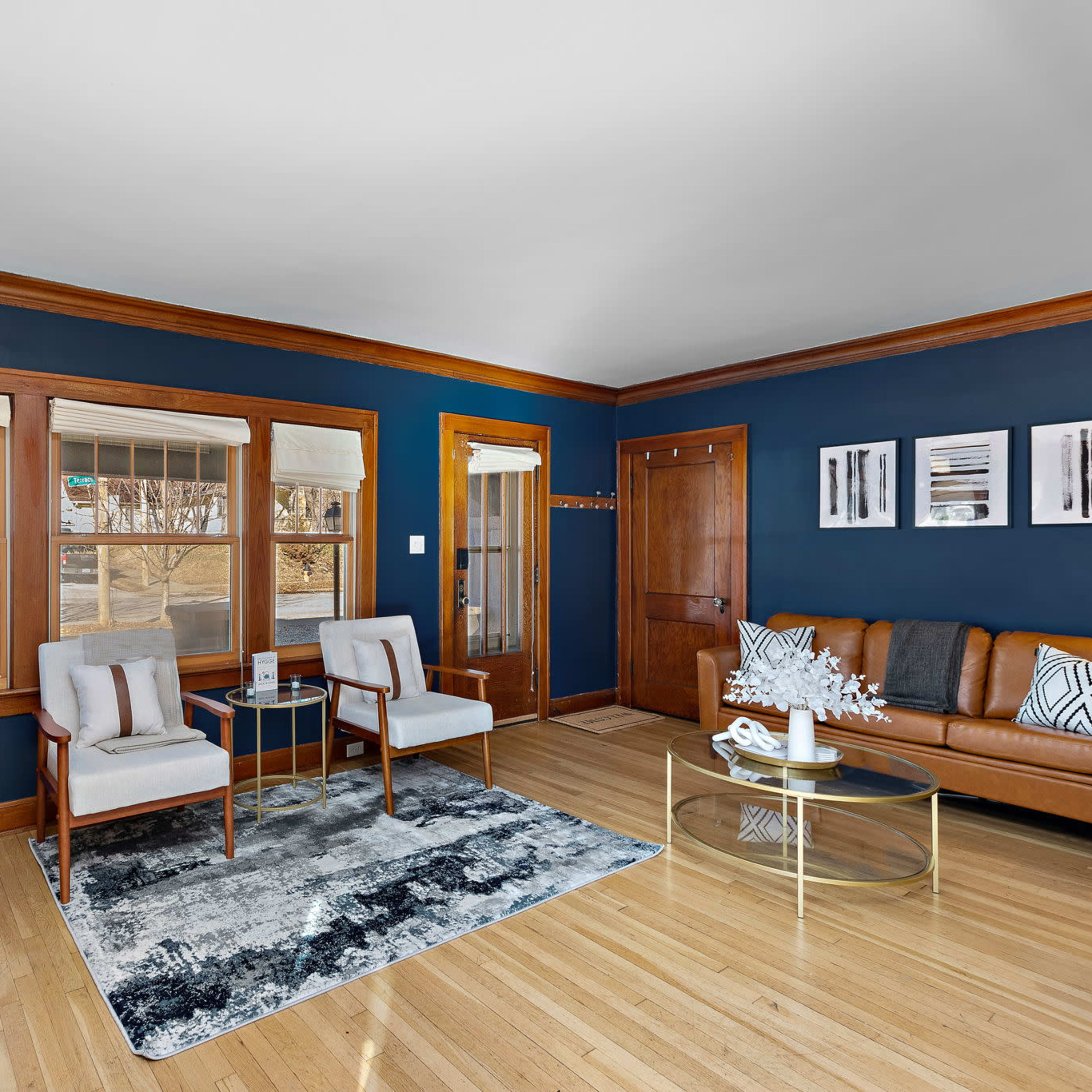 A living room with a brown leather sofa, two upholstered chairs, a glass coffee table, and large windows that provide natural light.