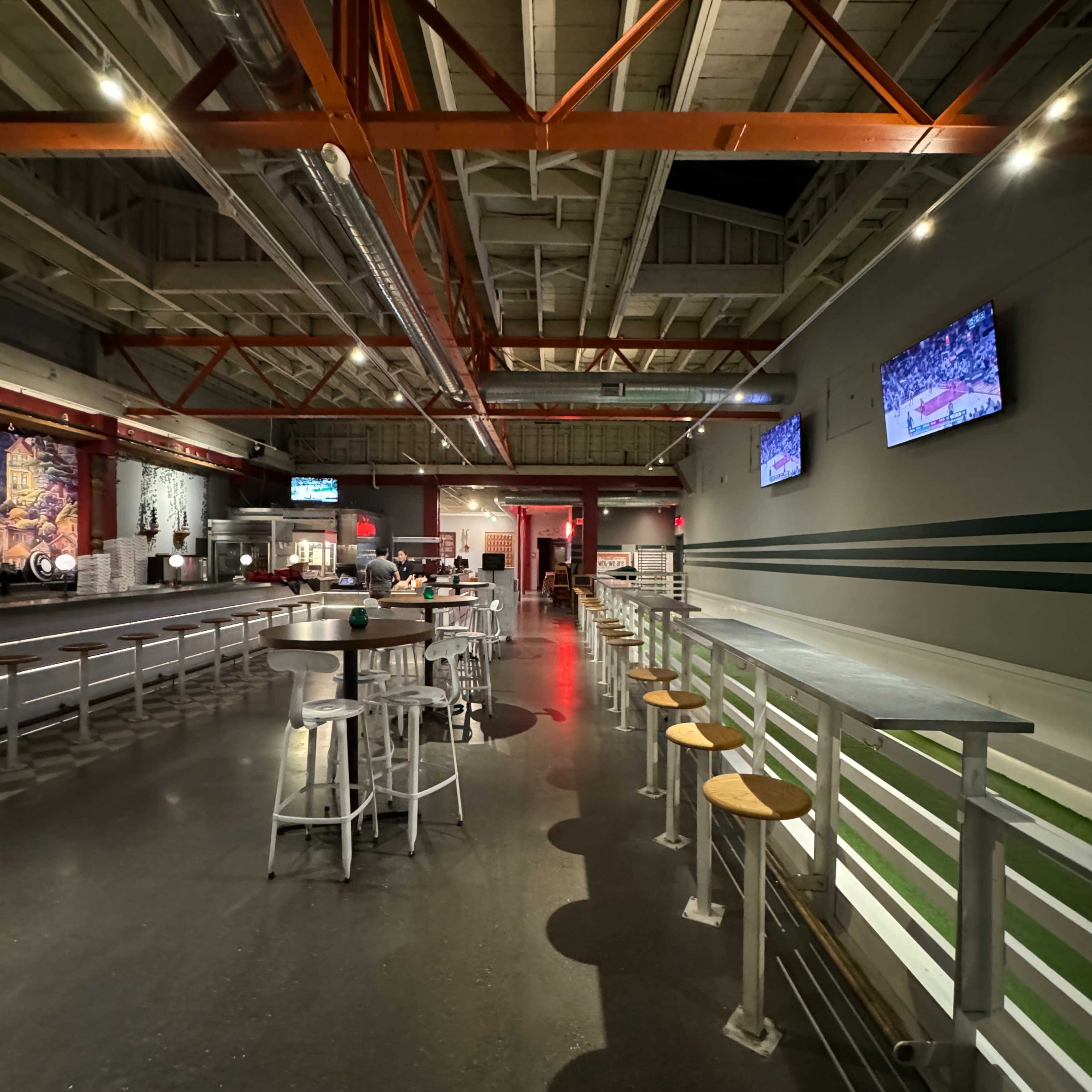 An empty bar area with high tables and stools, illuminated by overhead lighting and featuring multiple screens on the walls.