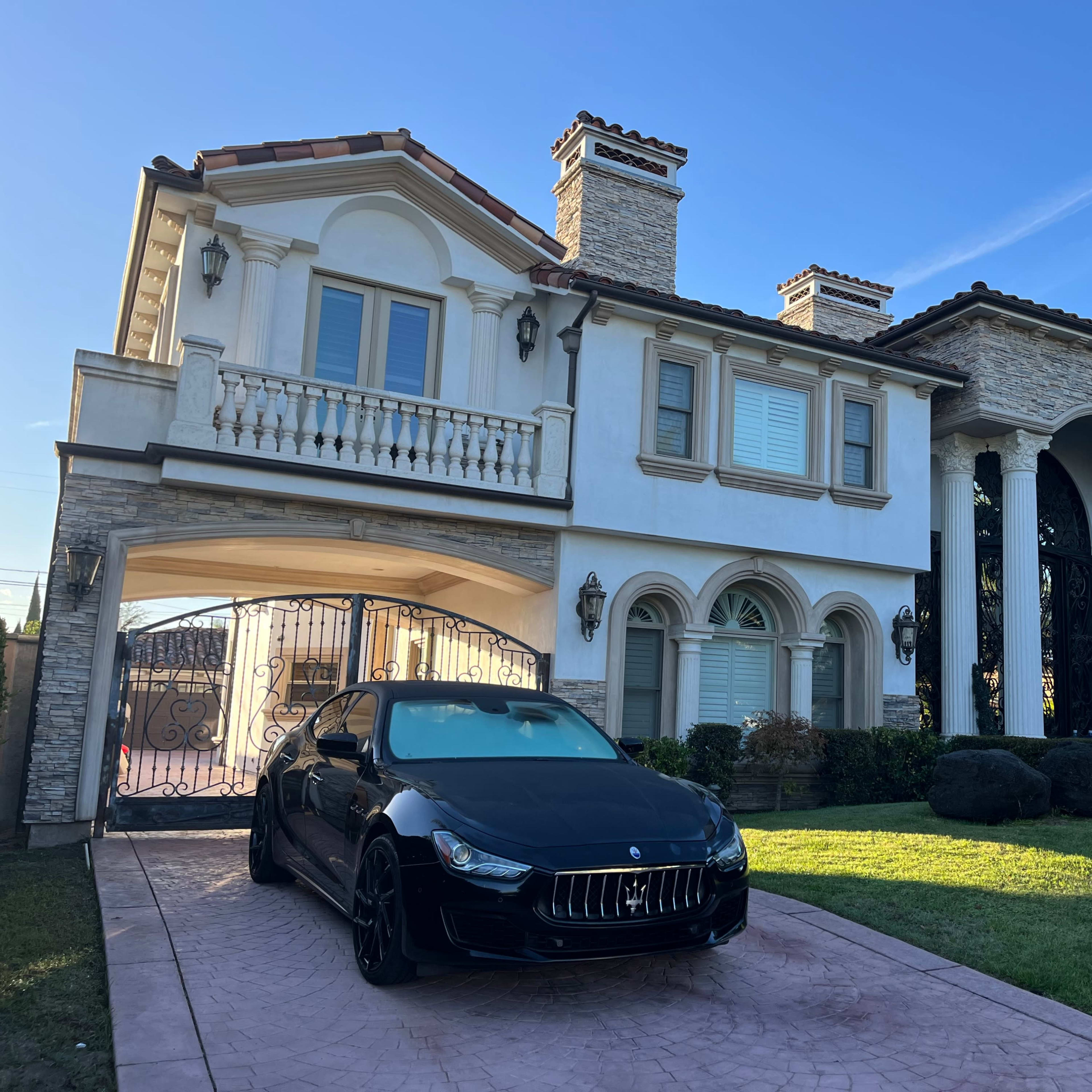 A large two-story house features a stone exterior, balconies, and a black car parked in the driveway.