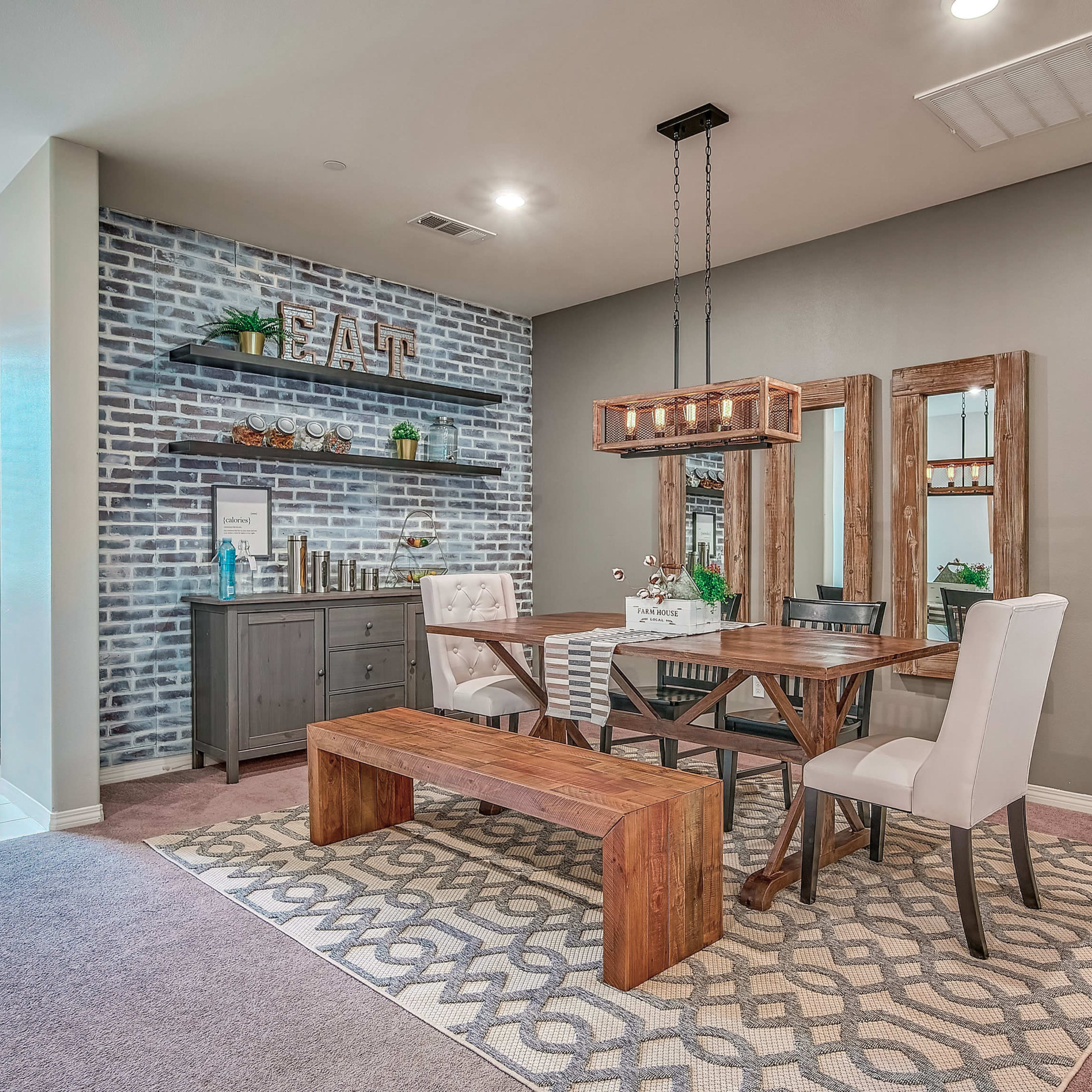 A dining area with a wooden table and chairs surrounded by a patterned rug, a gray sideboard, decorative mirrors, and a plant, against a backdrop of a brick feature wall.