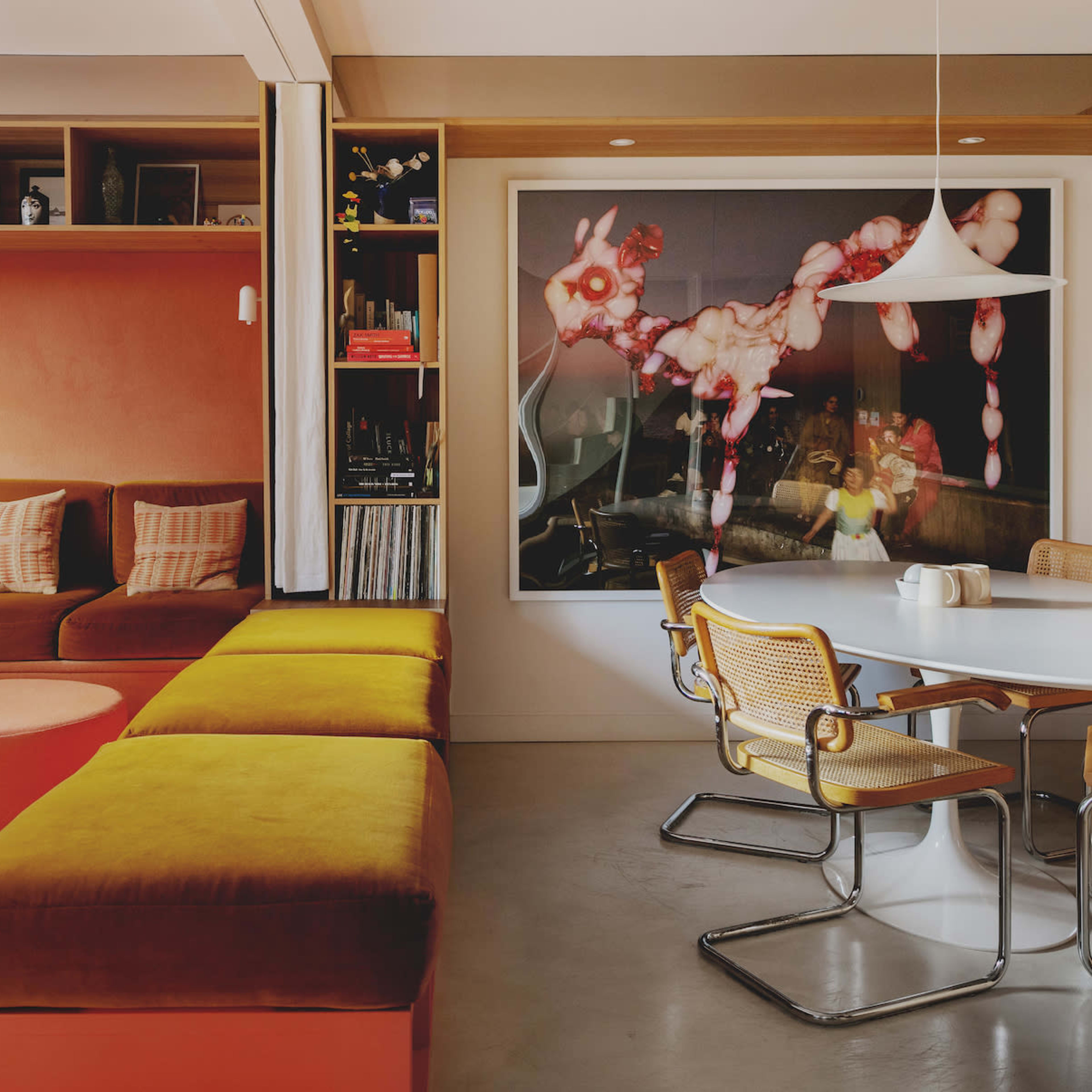 A cozy living space features a large, L-shaped sofa in earthy tones, a circular ottoman, and a round dining table surrounded by metal chairs, with a colorful artwork on the wall.