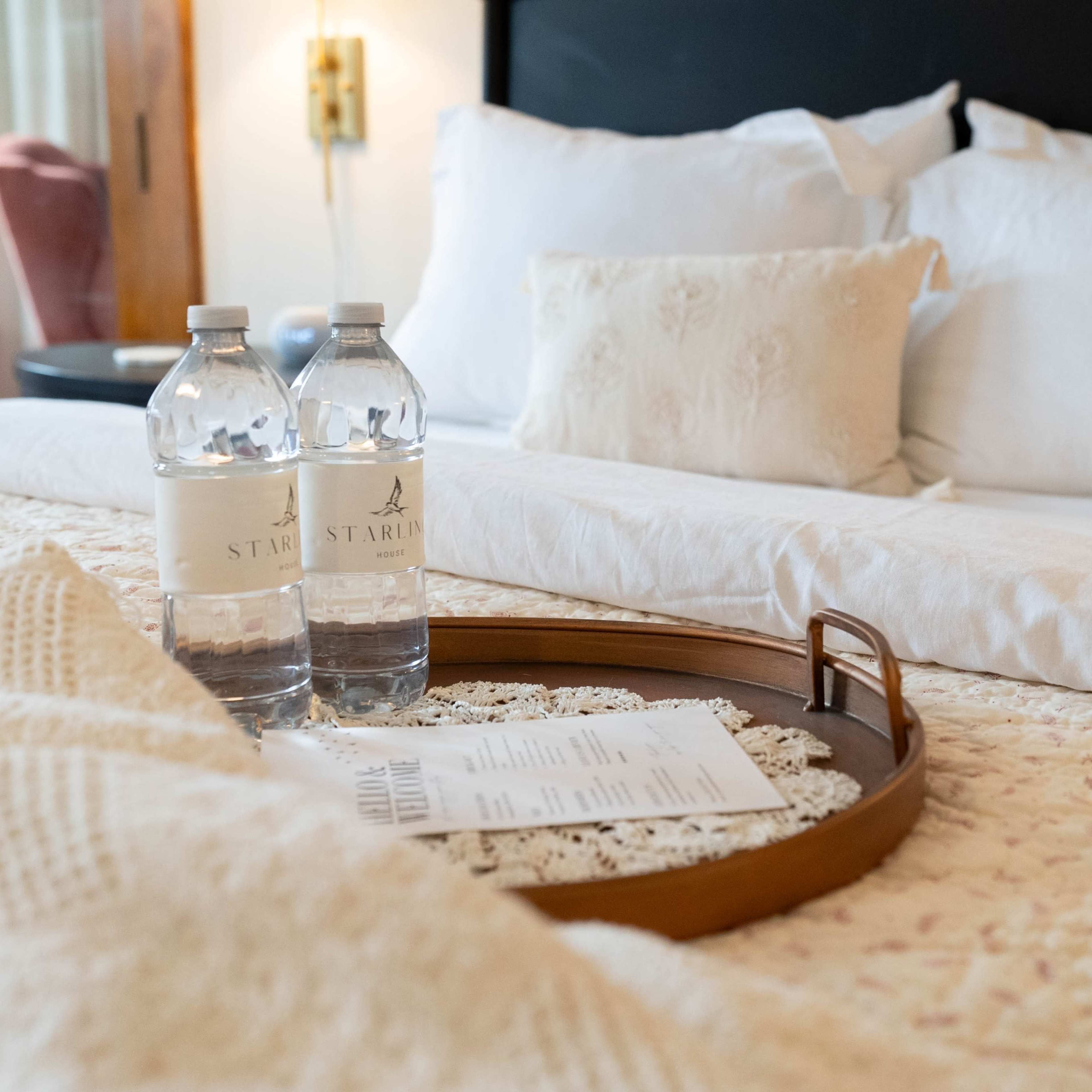 A neatly arranged hotel bed features two bottles of water on a wooden tray, along with a printed menu, set against a backdrop of white pillows.