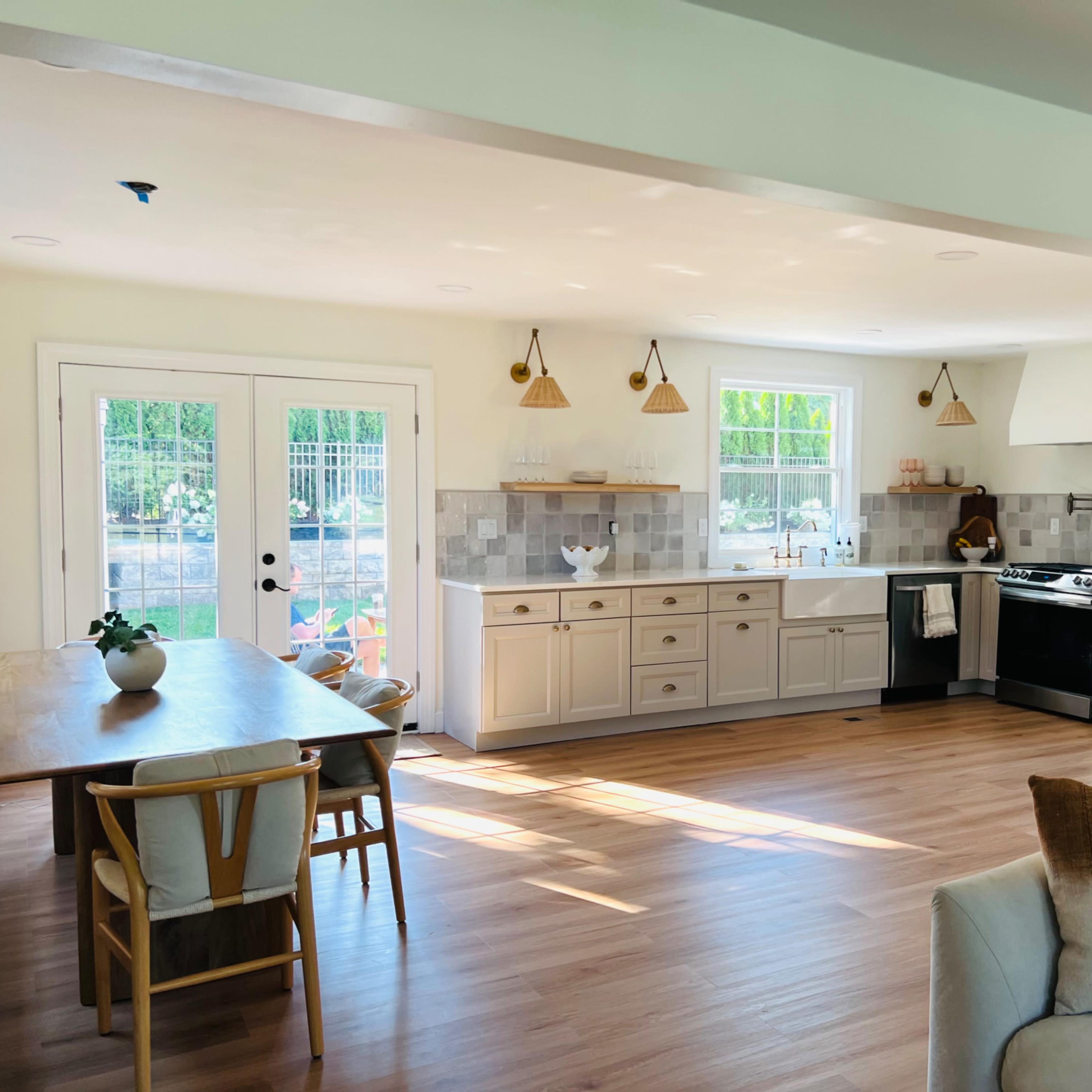 A bright and modern kitchen and dining area feature wooden flooring, white cabinetry, and large windows letting in natural light.