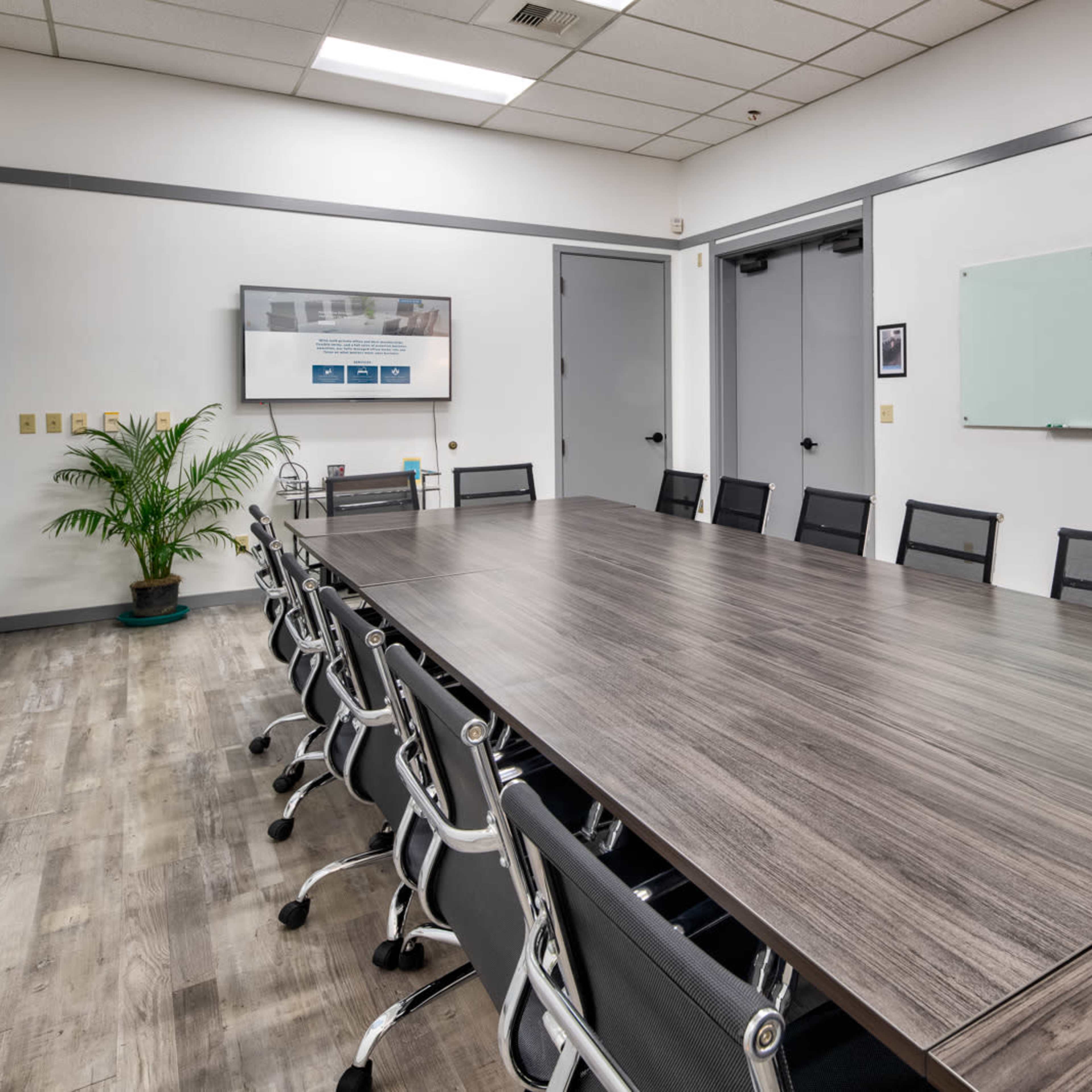 A large conference room with a long table surrounded by black mesh chairs, a whiteboard, and a wall-mounted screen.