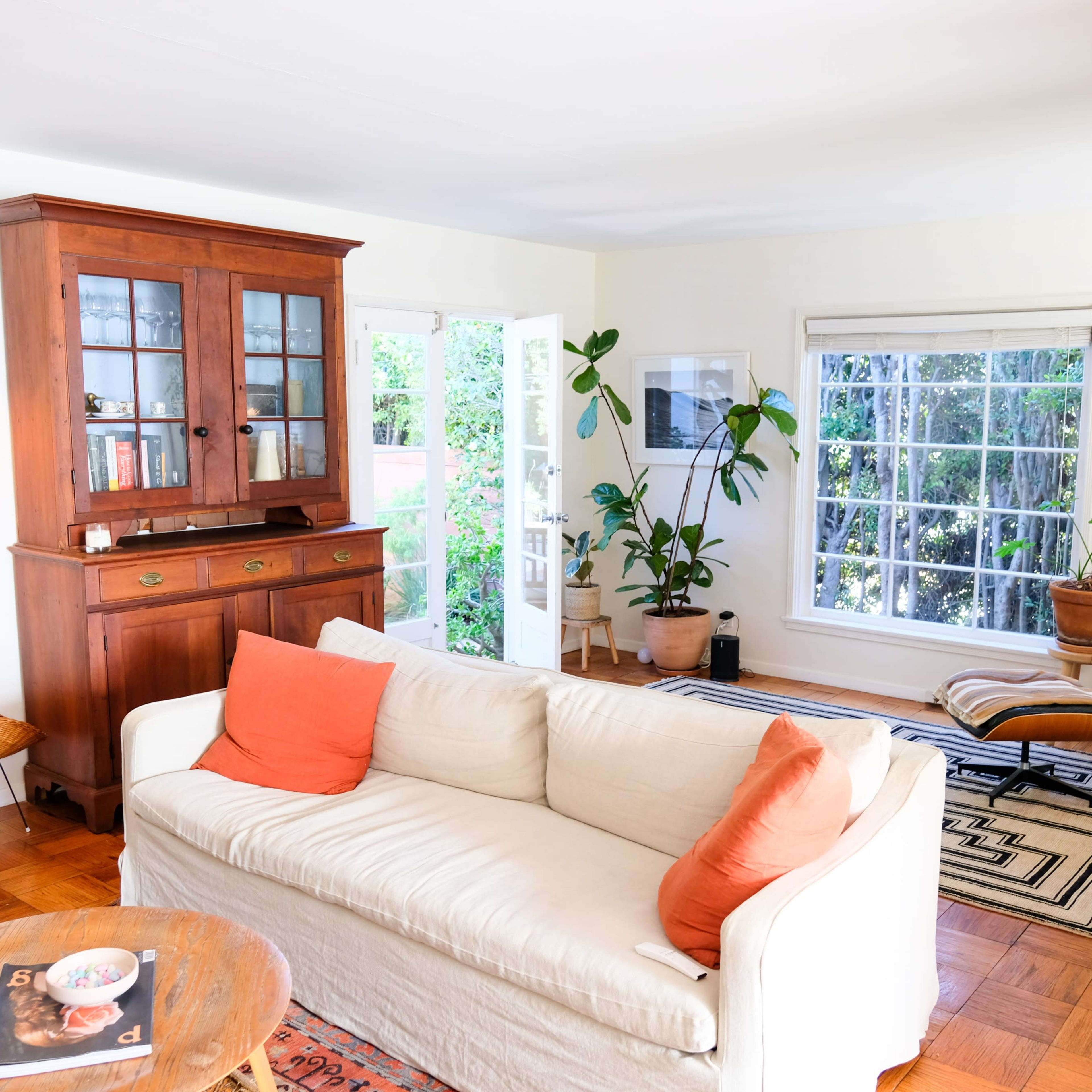 The living room features a beige sofa with orange cushions, a wooden cabinet, and large windows that provide natural light, surrounded by indoor plants.