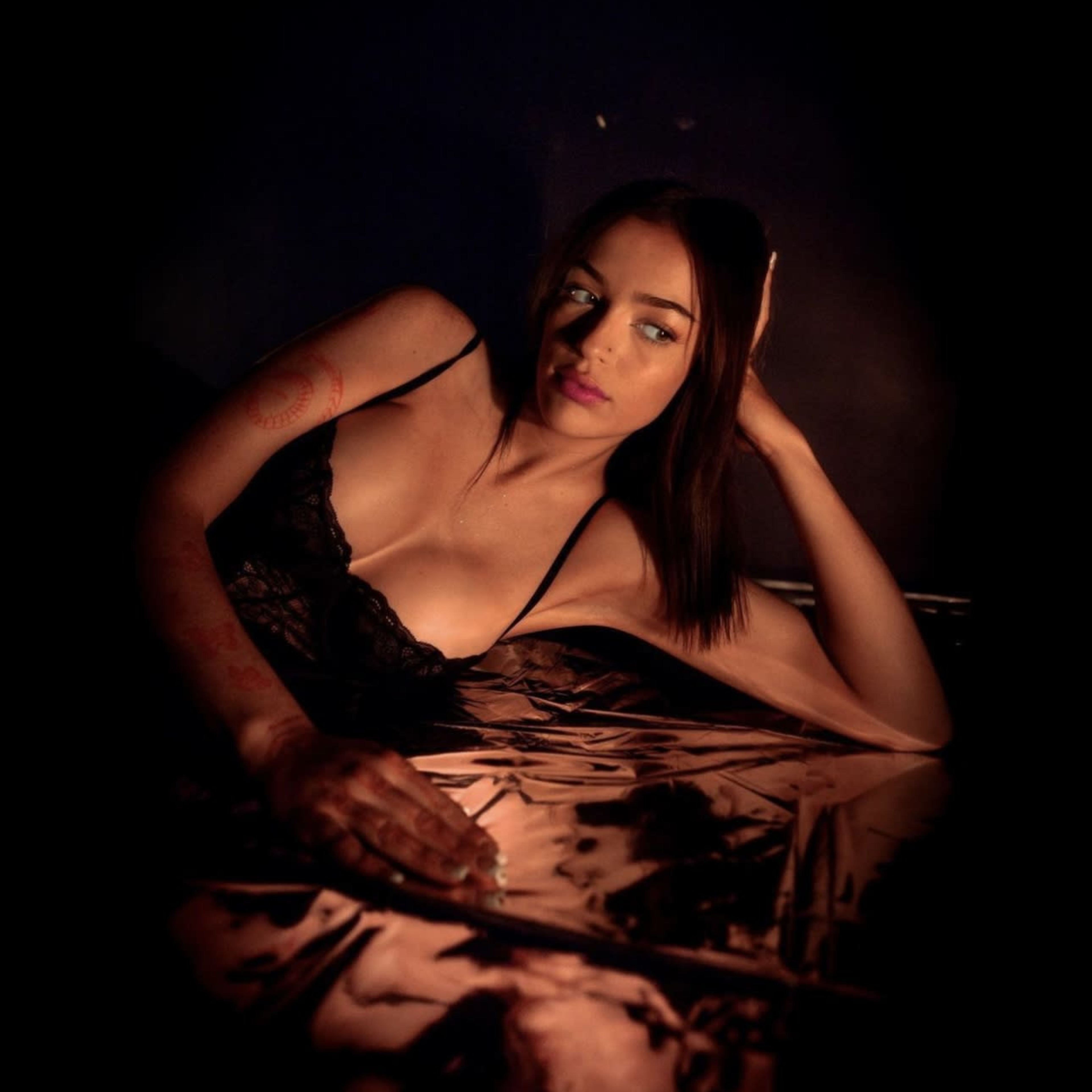 A woman in black lingerie reclines on a reflective surface in a dimly lit setting.