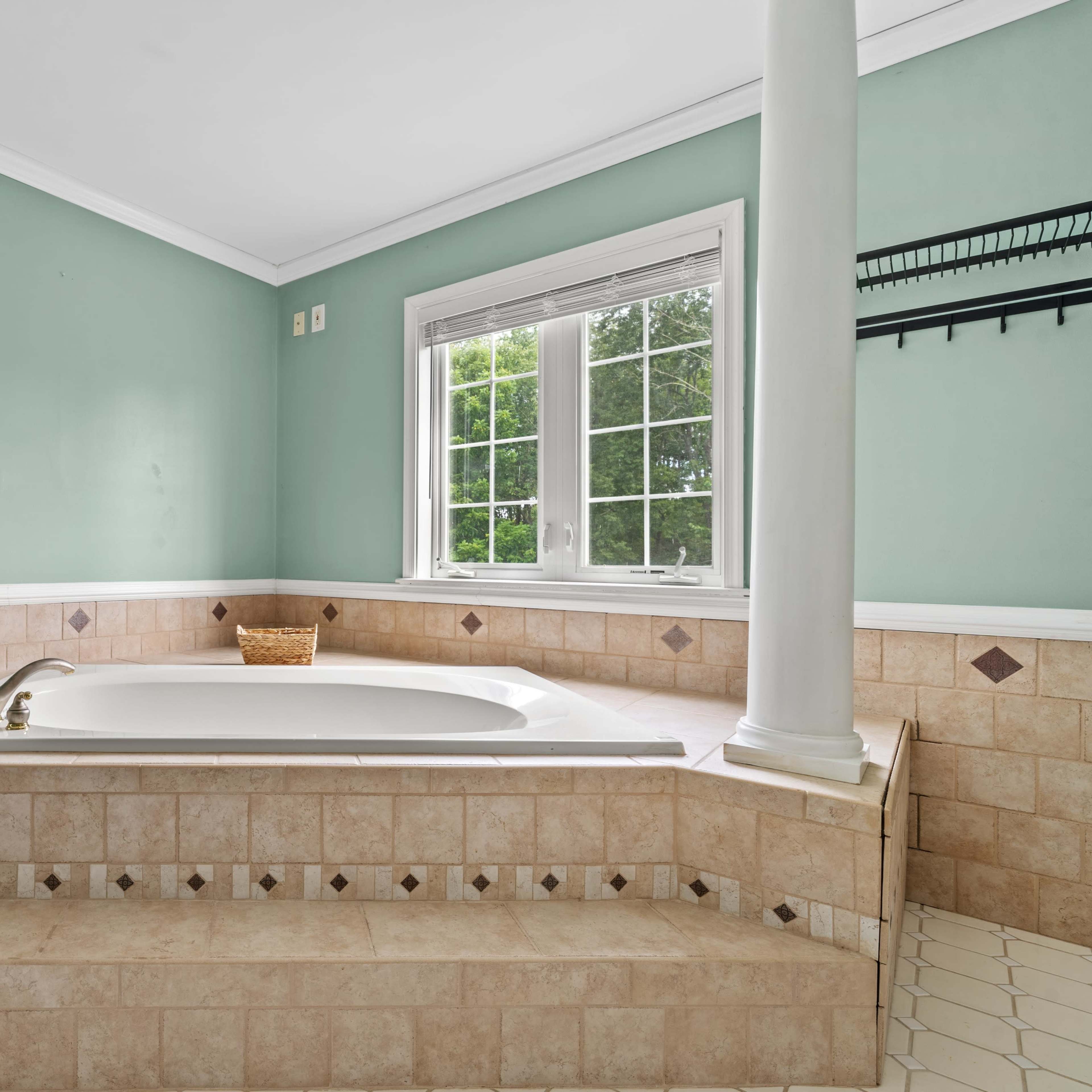 A large, tiled bathtub surrounded by two white columns in a bathroom with green walls and a window overlooking a garden.