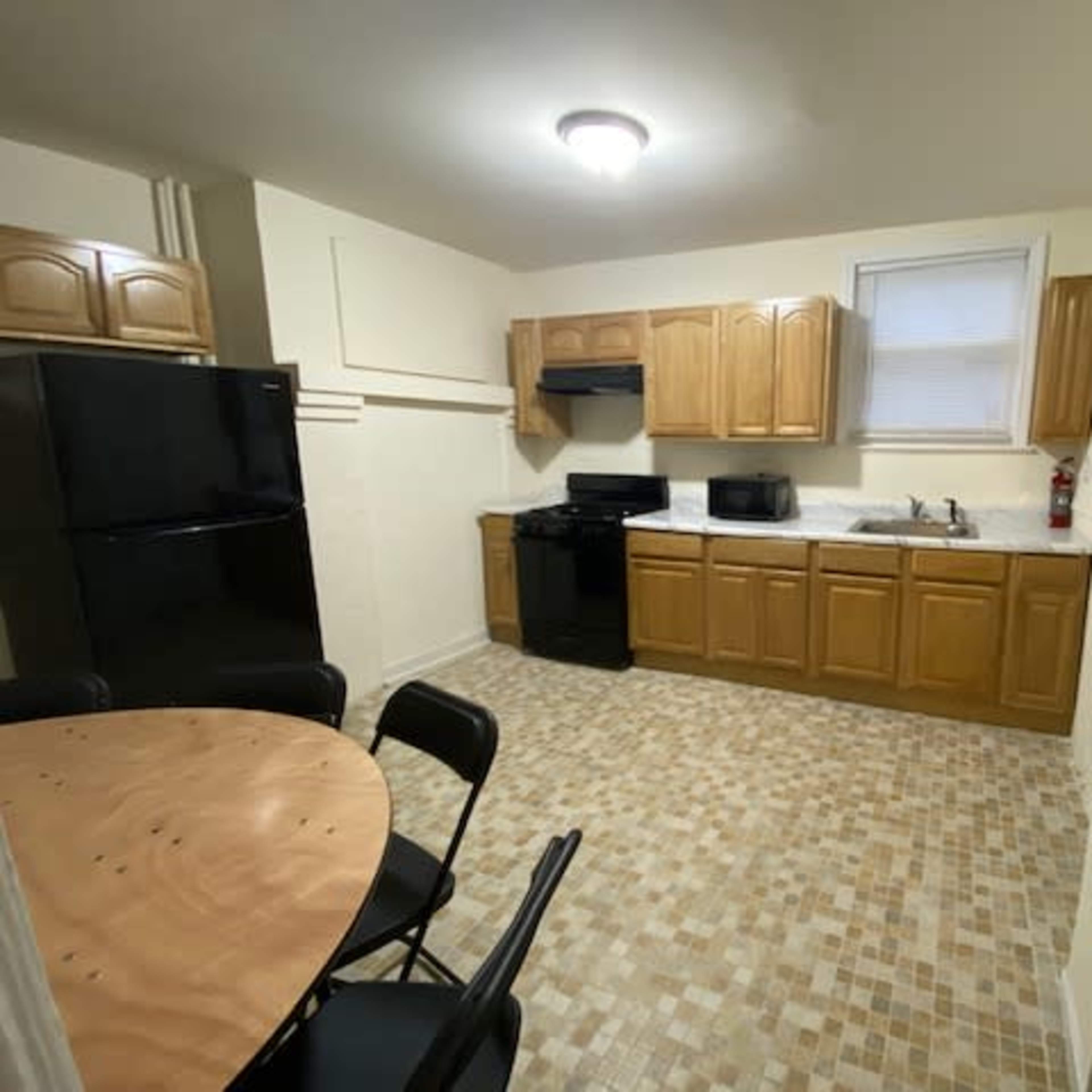 A small kitchen with wooden cabinets, a black refrigerator and stove, a sink, a microwave, and a round table with black chairs on a tiled floor.
