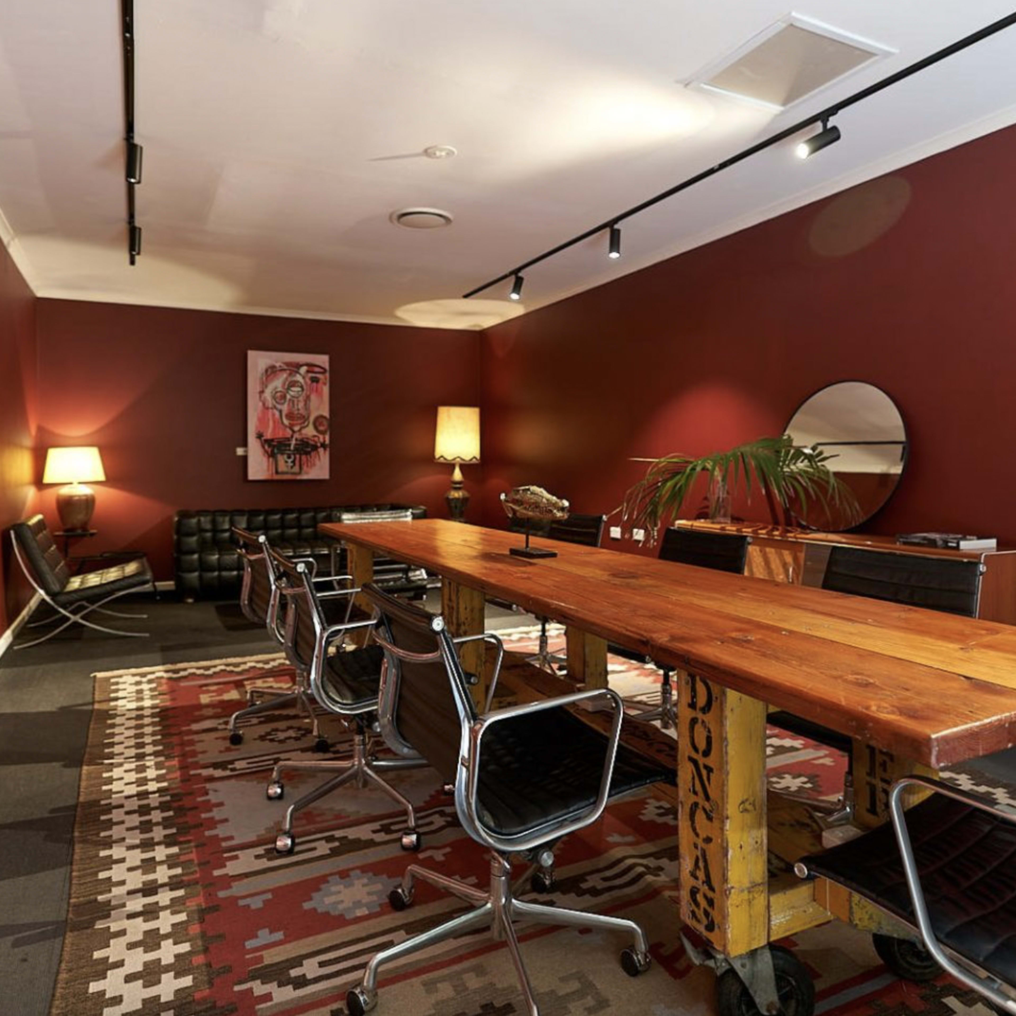 A modern meeting room features a long wooden table surrounded by black chairs, with a red wall, artwork, and a lamp in the background.