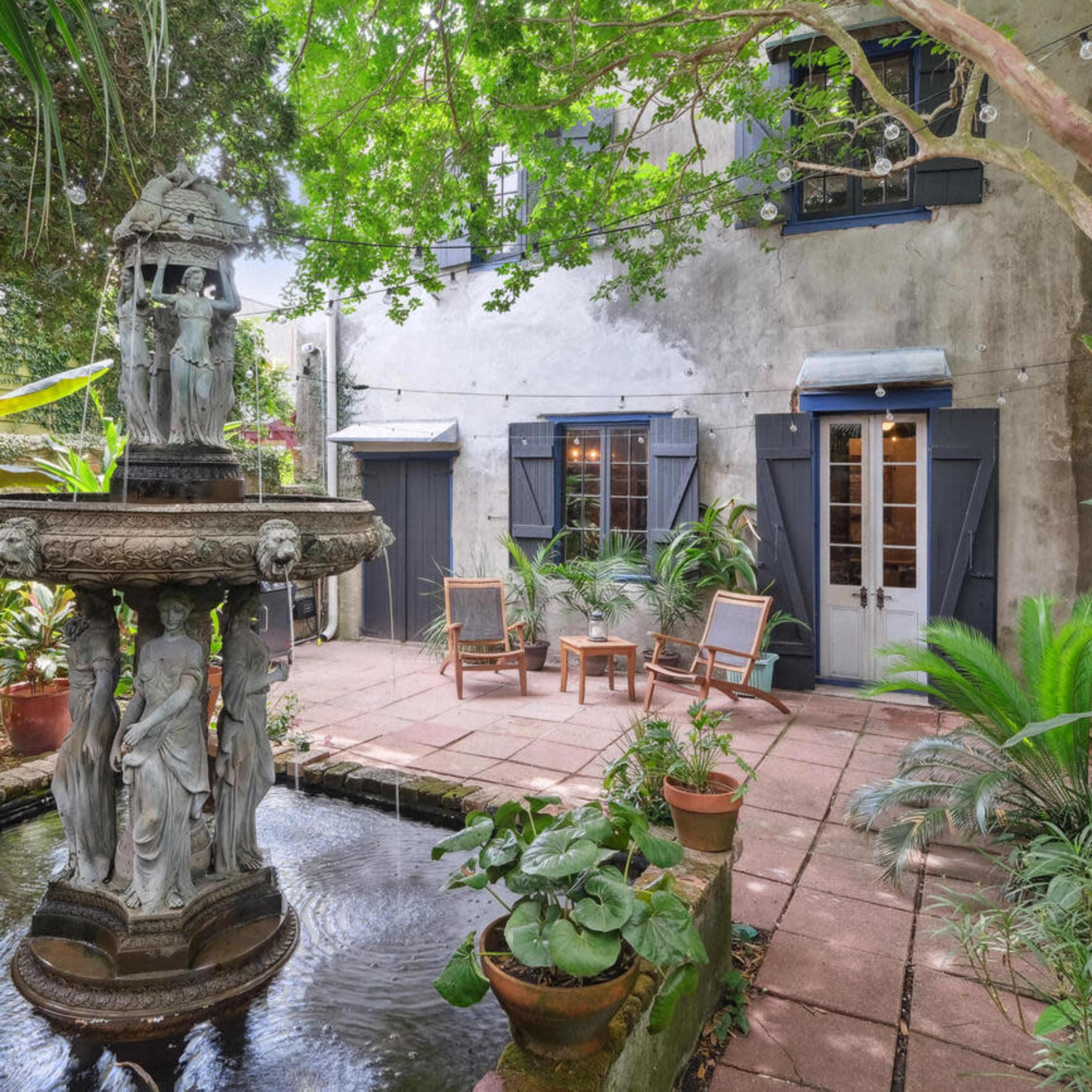 A serene courtyard featuring a central fountain surrounded by potted plants and chairs, with a weathered building in the background.
