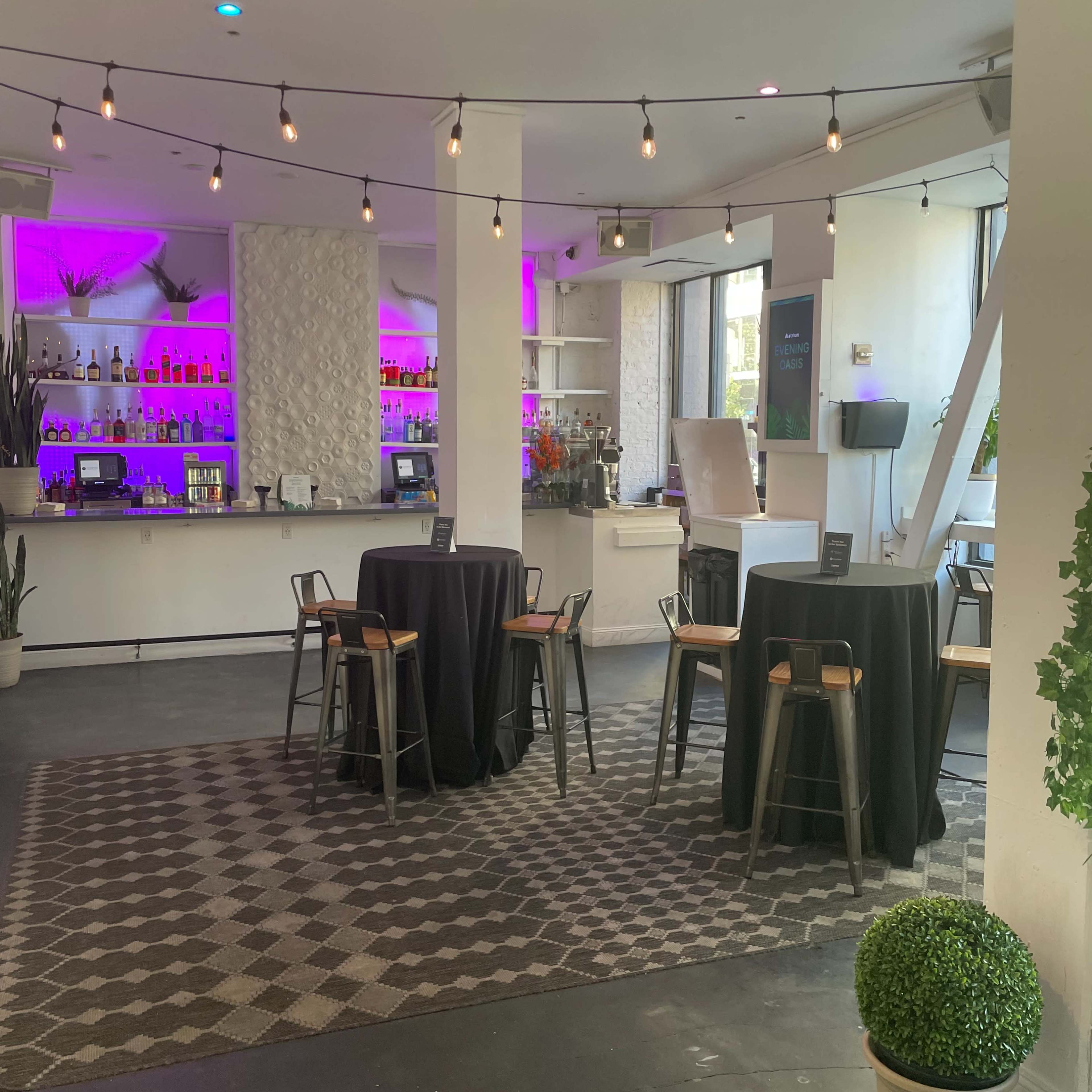 A modern bar area with a geometric-patterned rug, several high-top tables, and a backlit bar featuring bottles of various drinks.