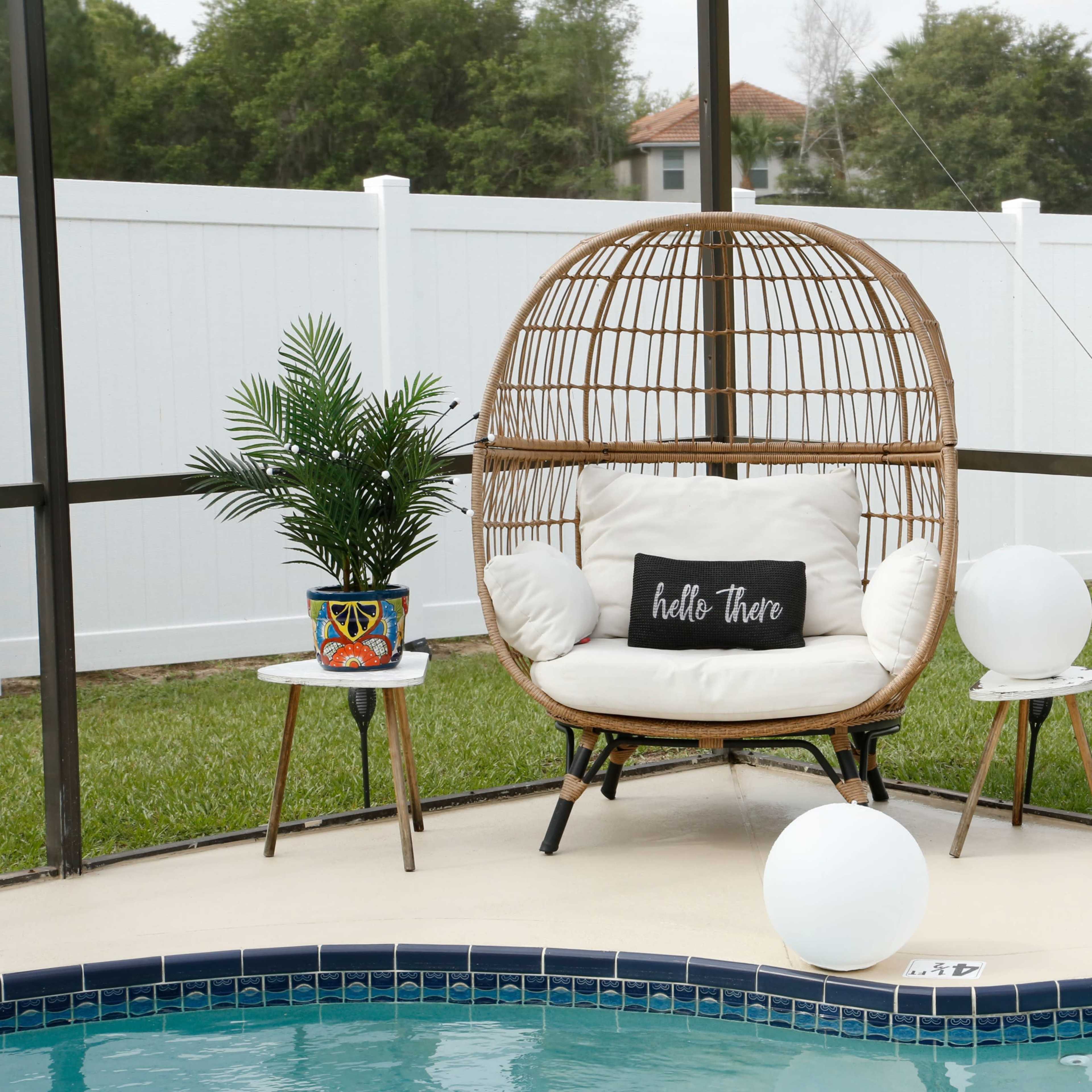 A wicker chair with a cushion and a decorative pillow sits beside a pool, accompanied by a potted plant and two small tables.