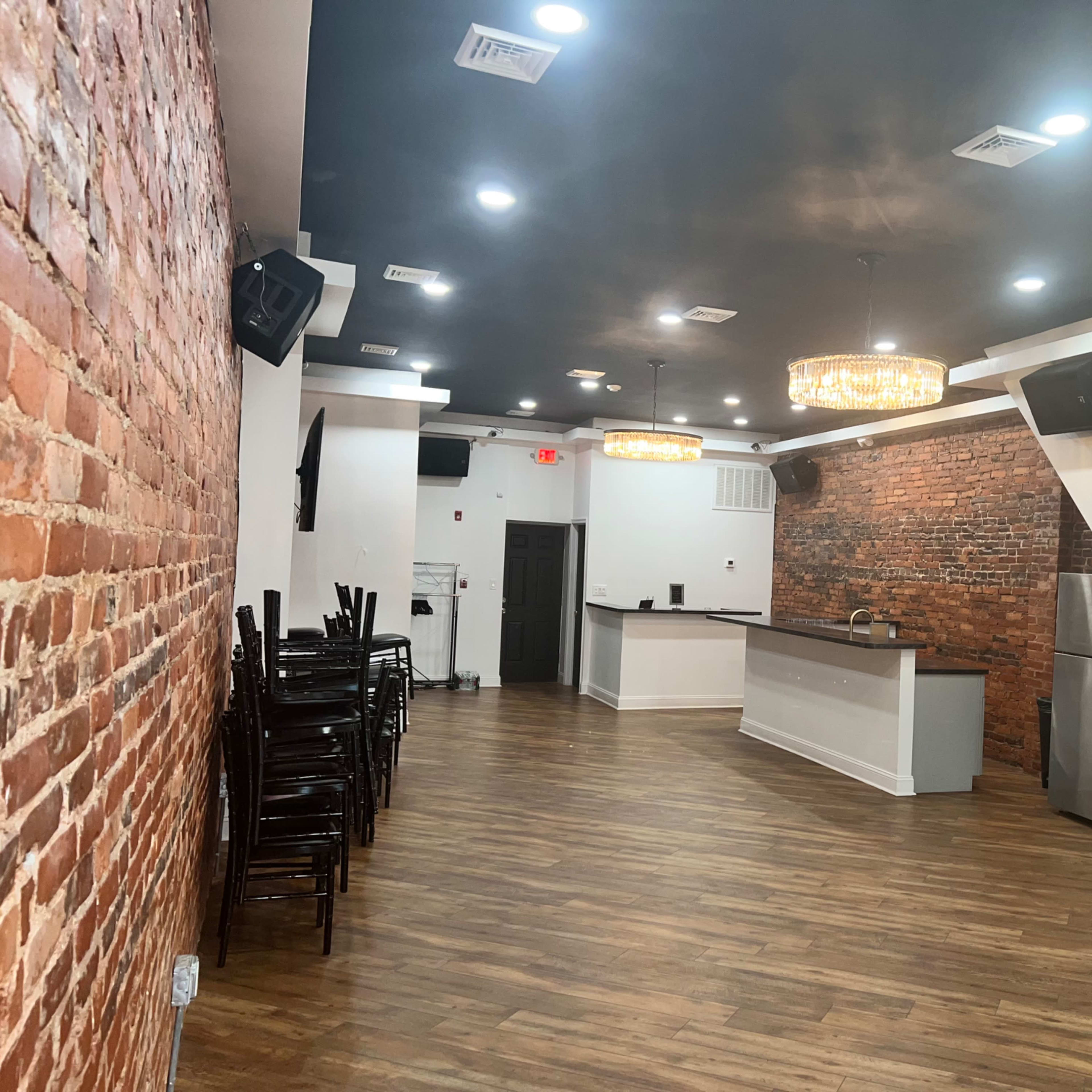 A spacious interior featuring exposed brick walls, a bar counter, and wooden flooring with stacked chairs along one side.