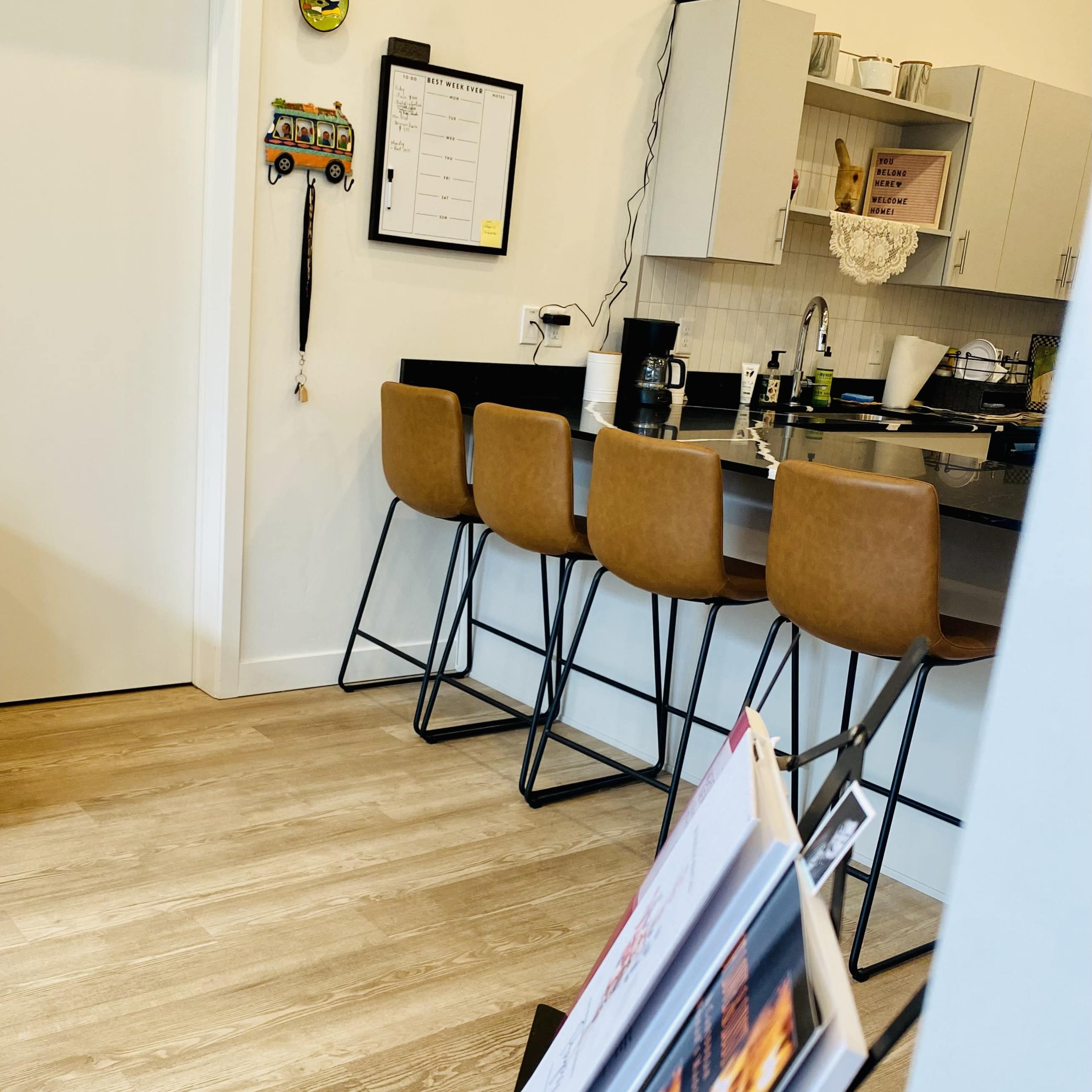 A modern kitchen with a bar counter and four stools, a wall-mounted calendar, and a magazine rack in the foreground.