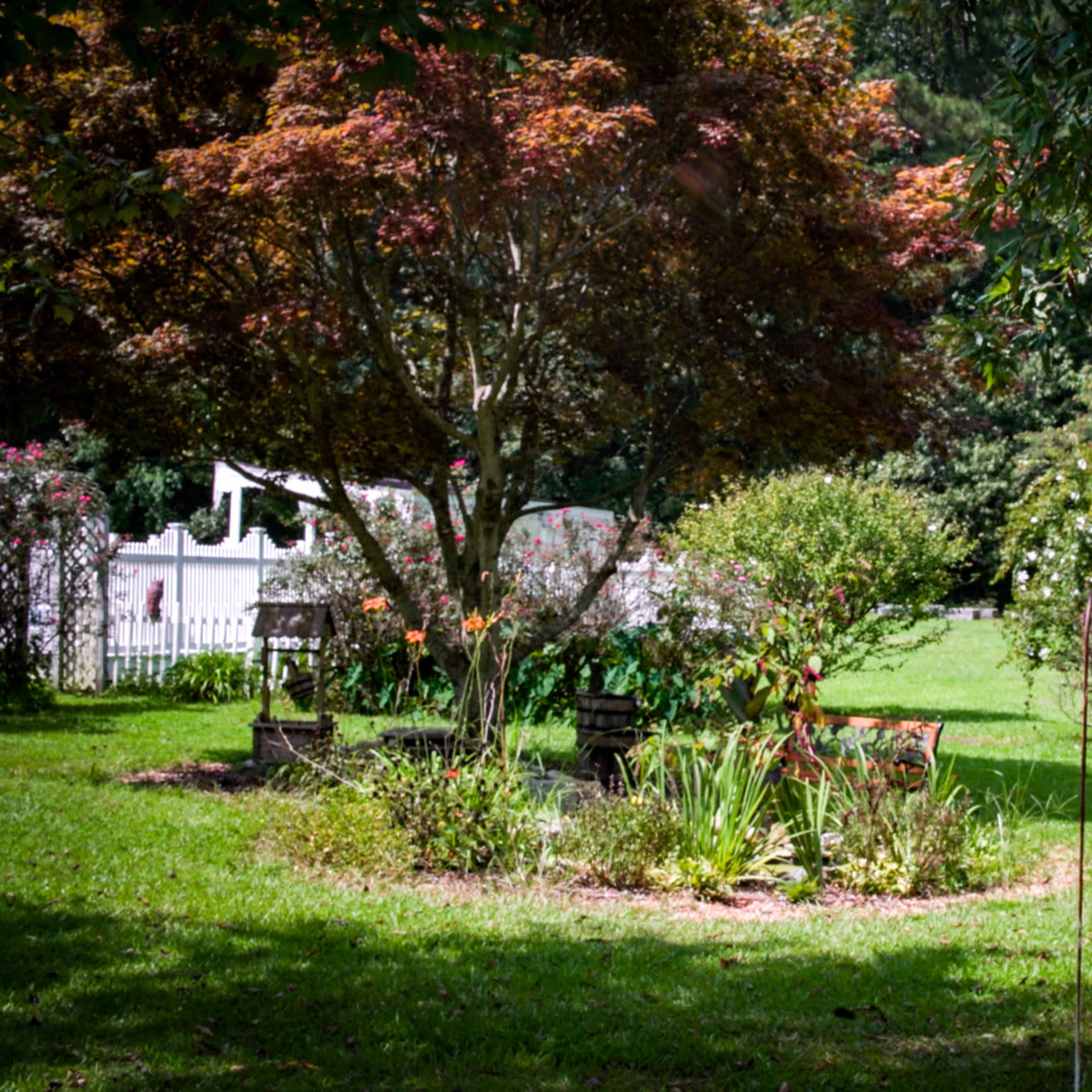 A well-maintained garden features colorful flowers, shrubs, and a small tree, surrounded by a white picket fence.