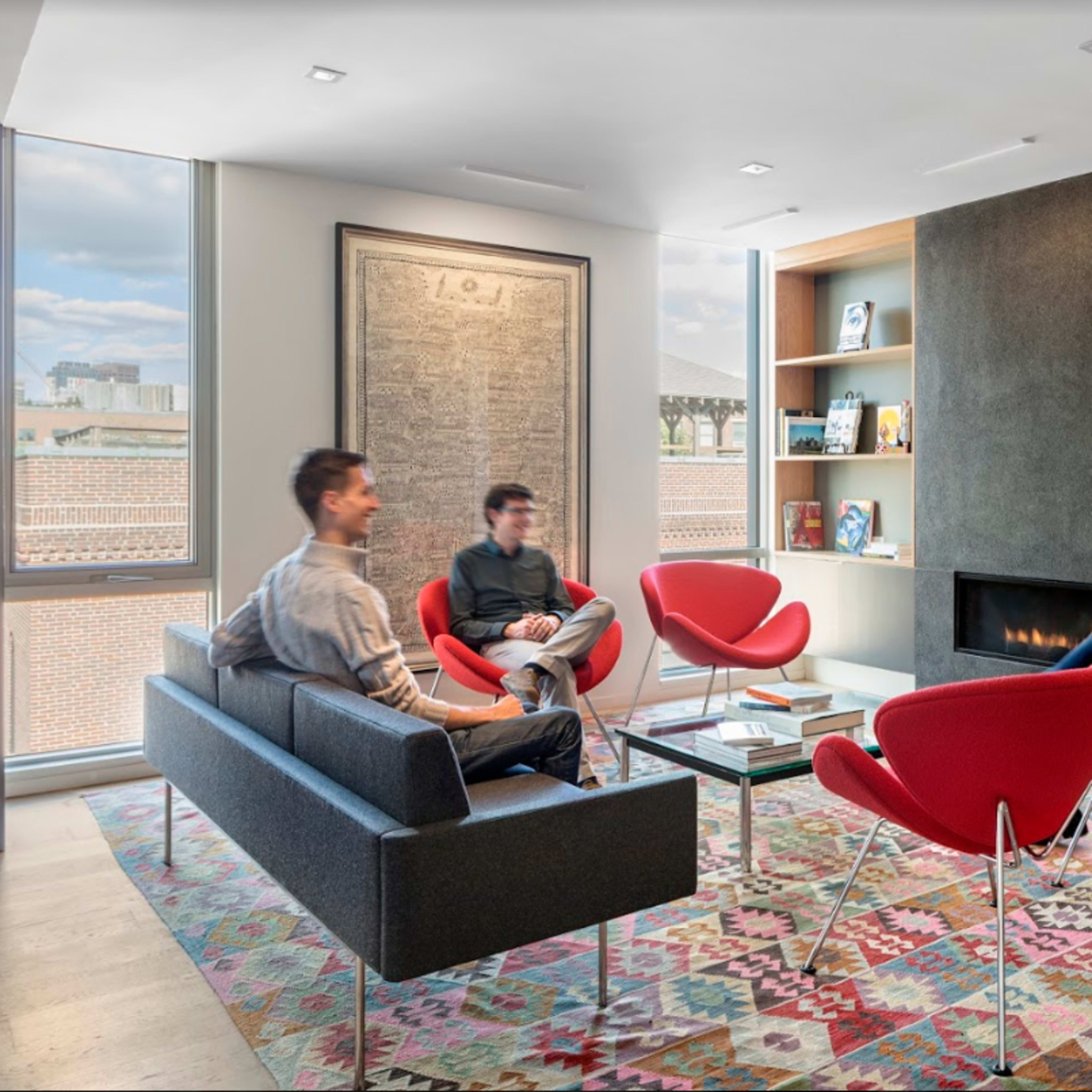 A modern living room features four people seated in red chairs and a gray sofa, with a fireplace and bookshelves in the background.