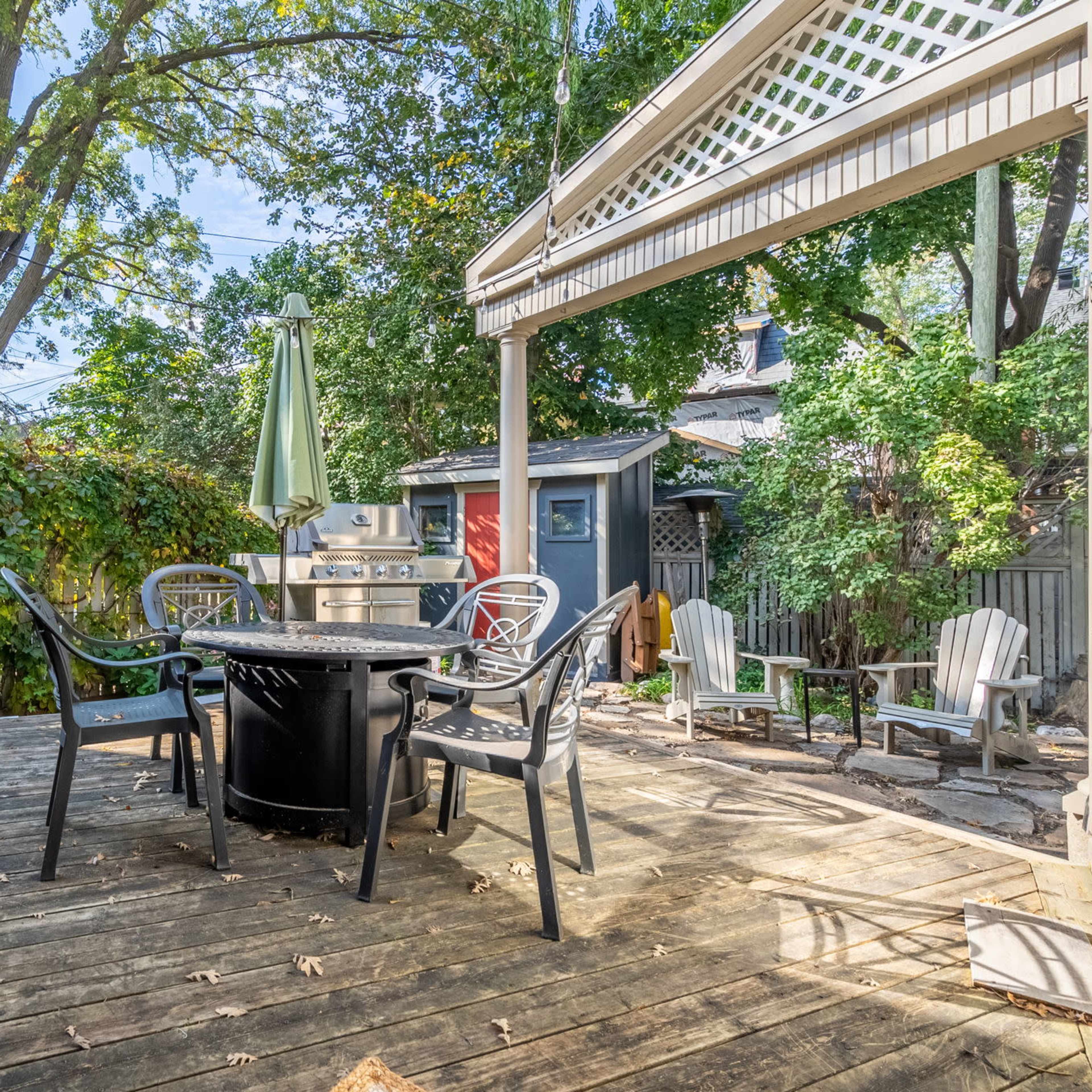 A wooden deck with a circular table and chairs, surrounded by greenery and a small shed in the background.