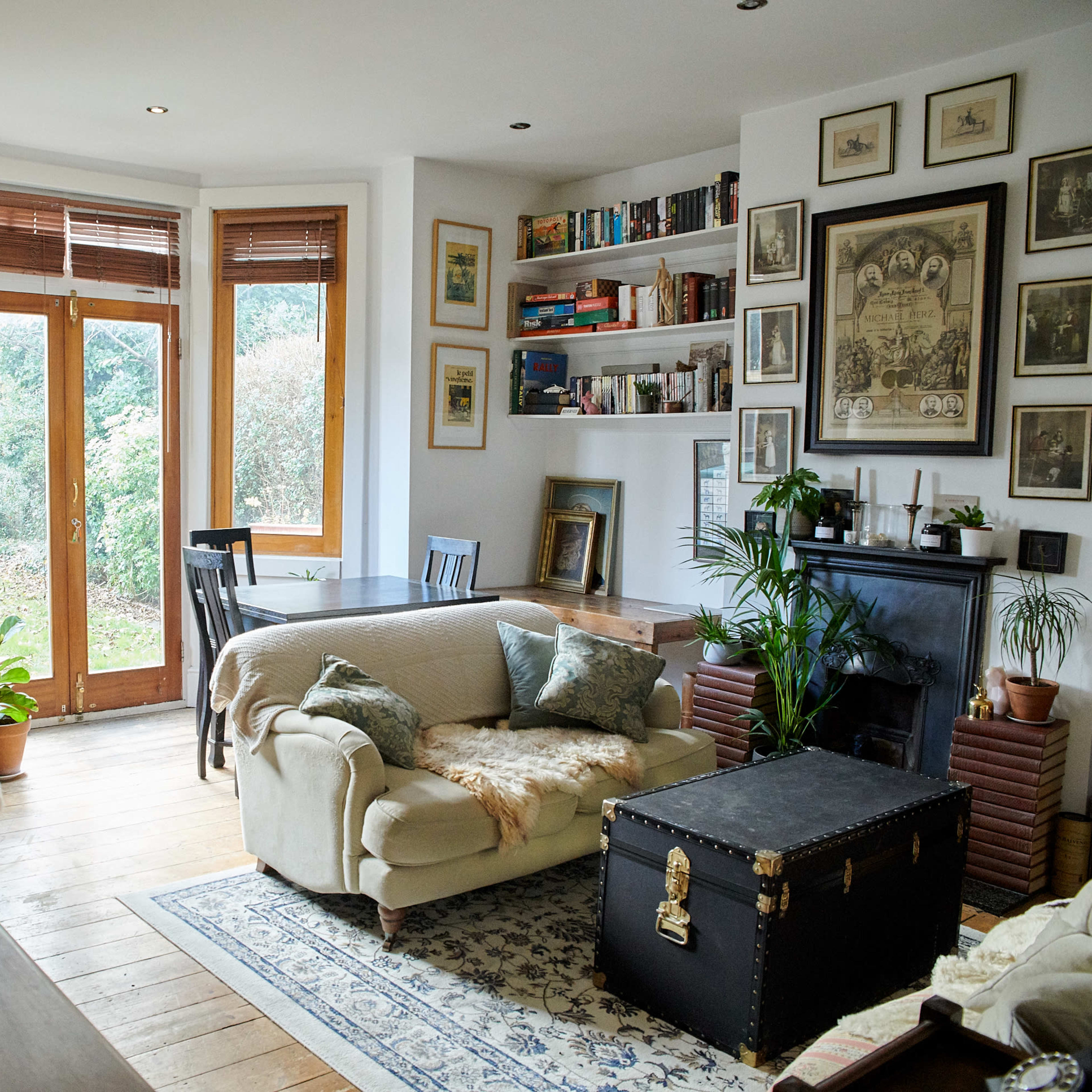 A cozy living room features a mix of bookshelves, a fireplace, and large windows overlooking a garden.