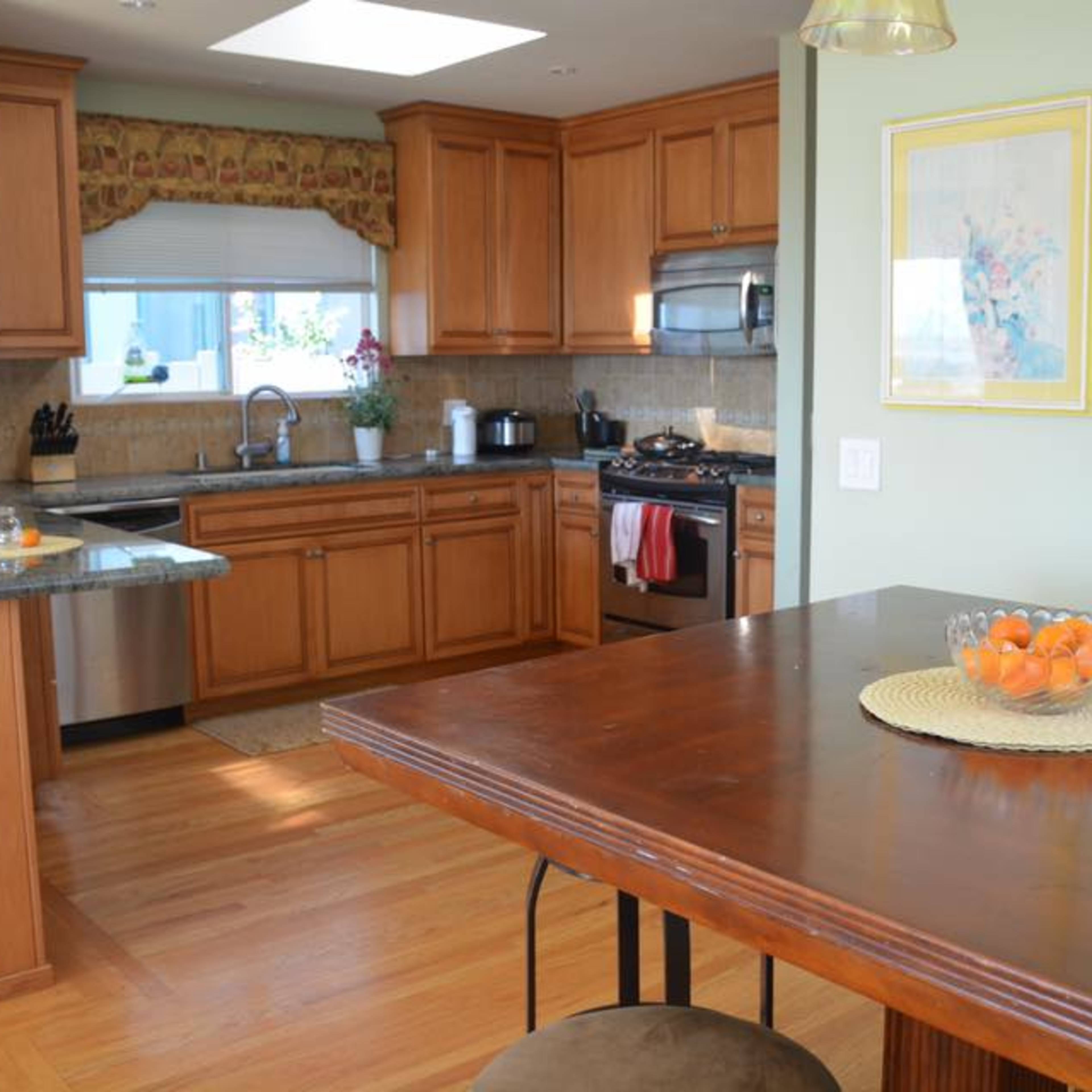 A well-lit kitchen featuring wooden cabinets, a granite countertop, and a dining table set with a bowl of oranges.