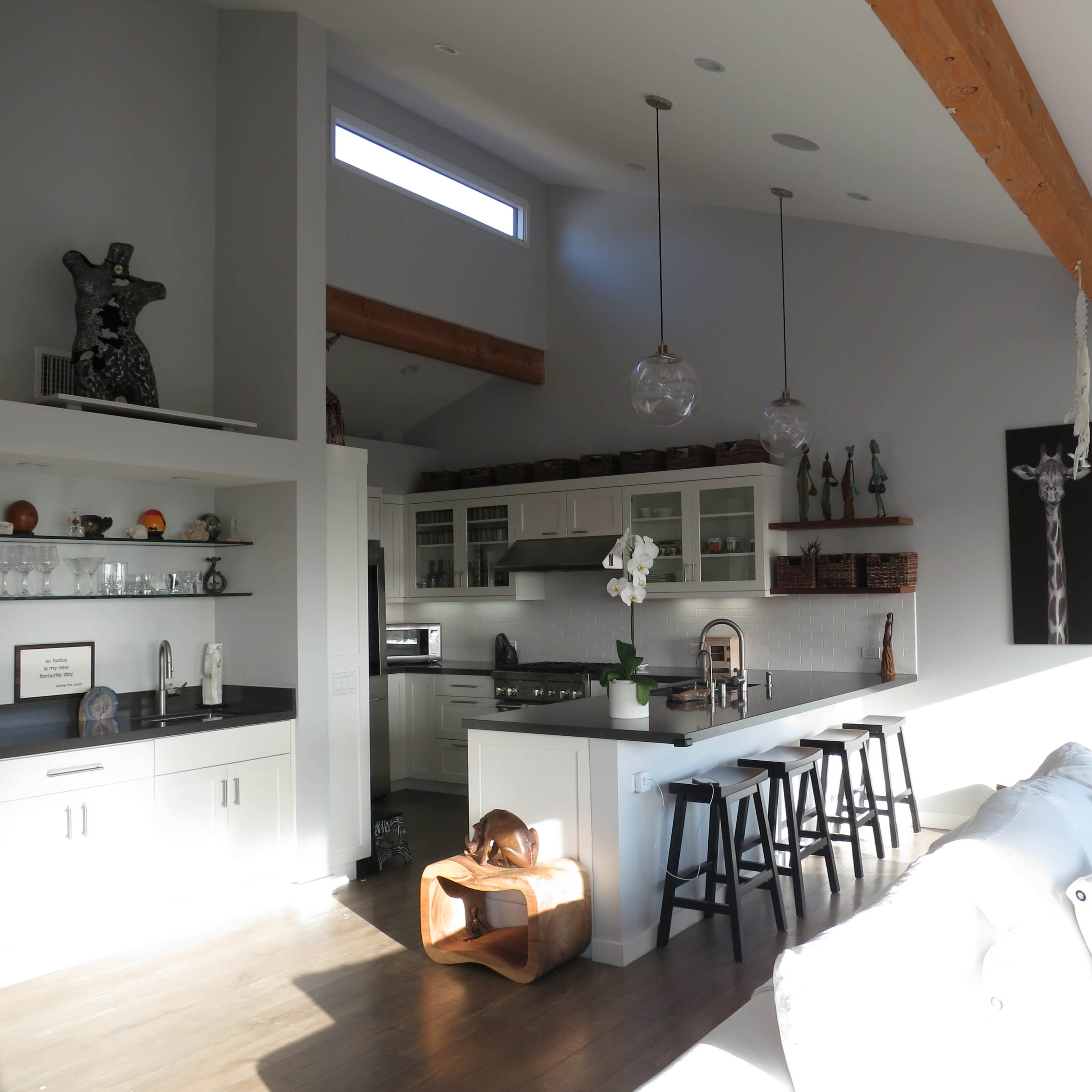 A modern kitchen and living area featuring a high ceiling, wooden beams, white cabinetry, and a sofa facing the kitchen island.