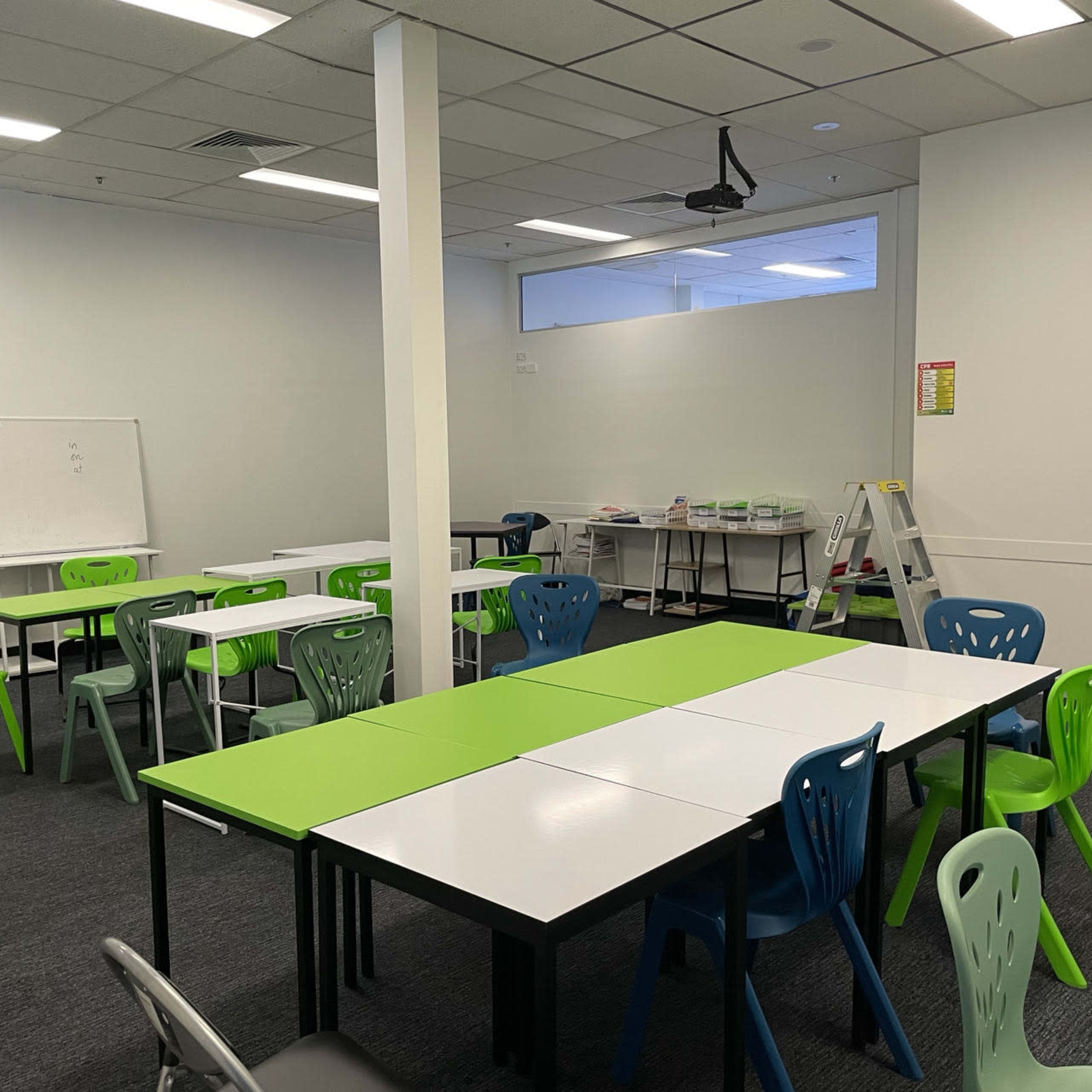 A classroom setting features multiple tables with green and white surfaces, accompanied by colorful chairs and a whiteboard, all arranged in a spacious area.
