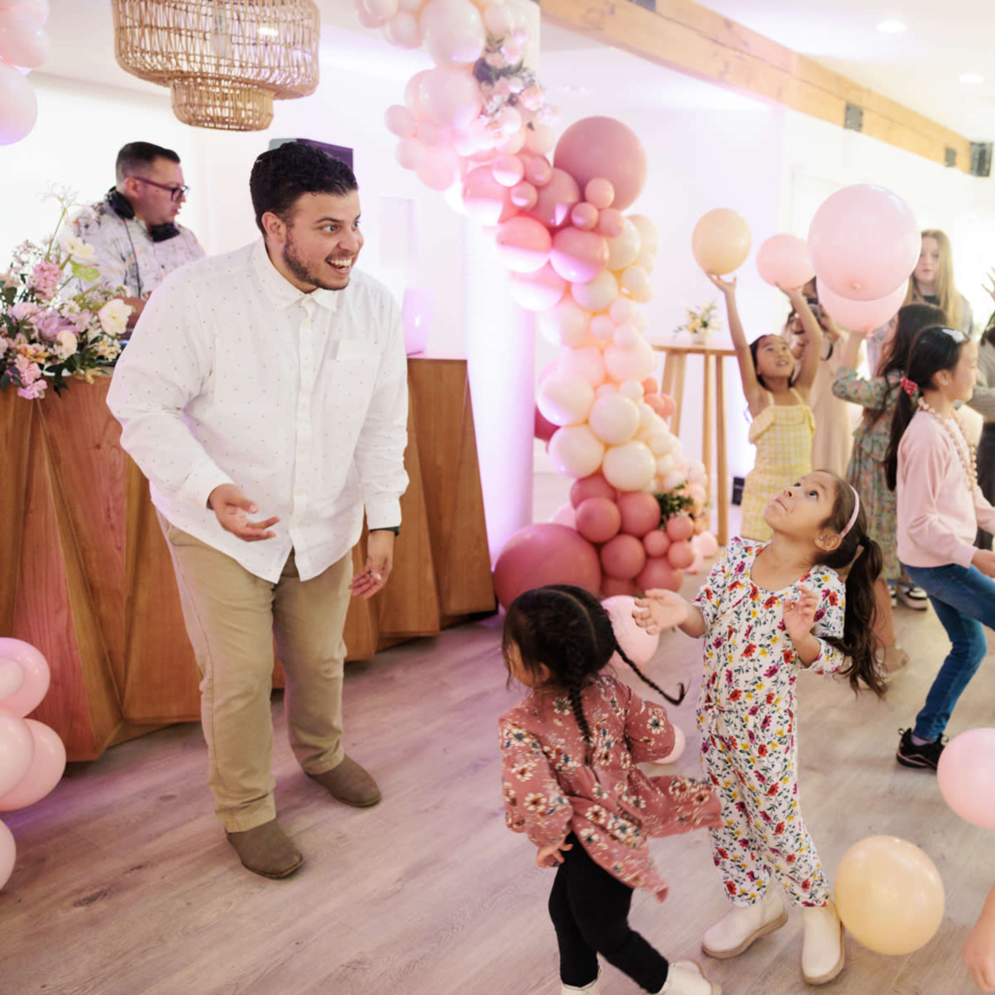 A group of children and adults dance joyfully at a celebration, surrounded by colorful balloons and decorations.