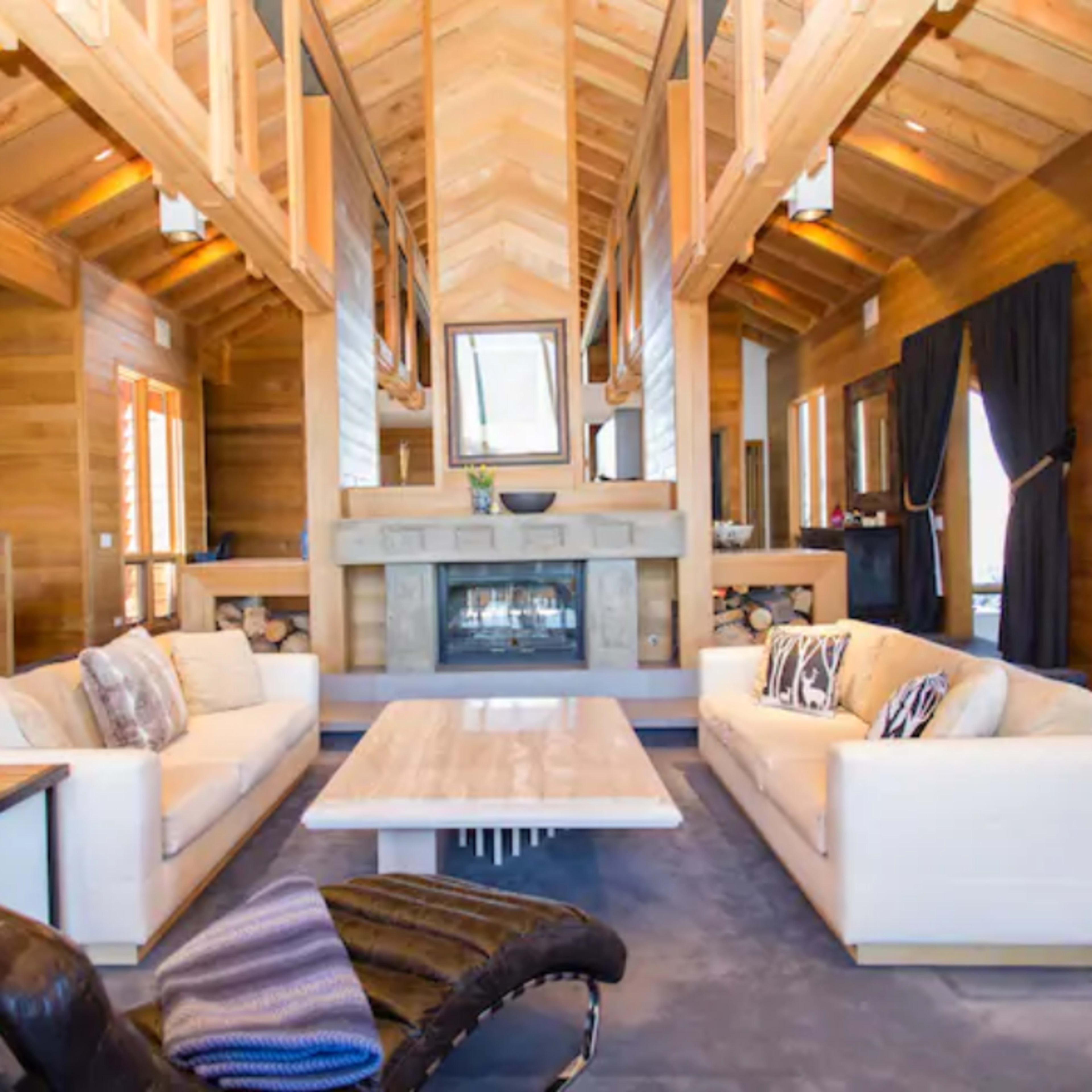 The living room features a high wooden ceiling, two white sofas facing a stone fireplace, and a light-colored coffee table.