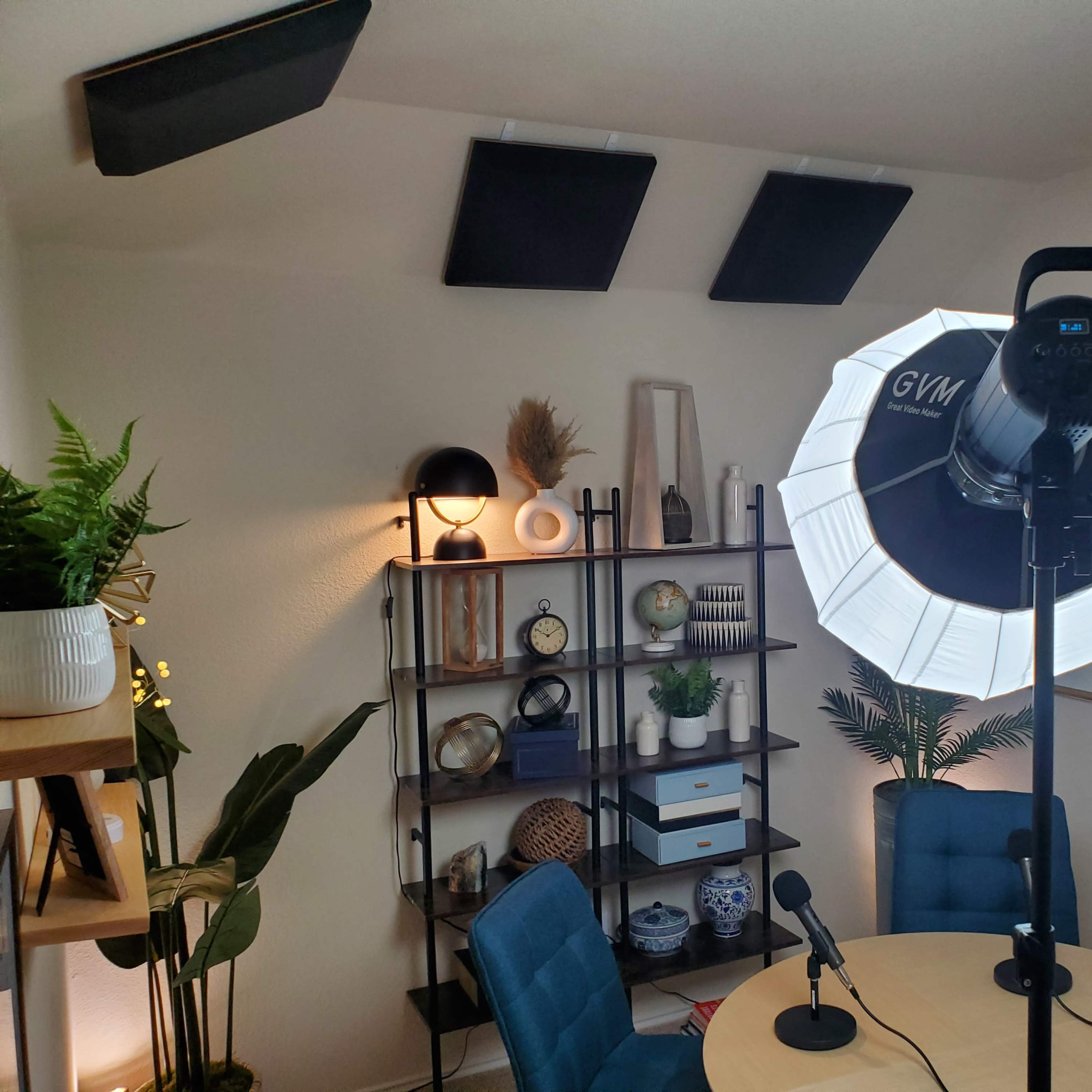 A well-lit room set up for a podcast or video recording, featuring a round table with two microphones, shelves with decorative items, and a large softbox light.
