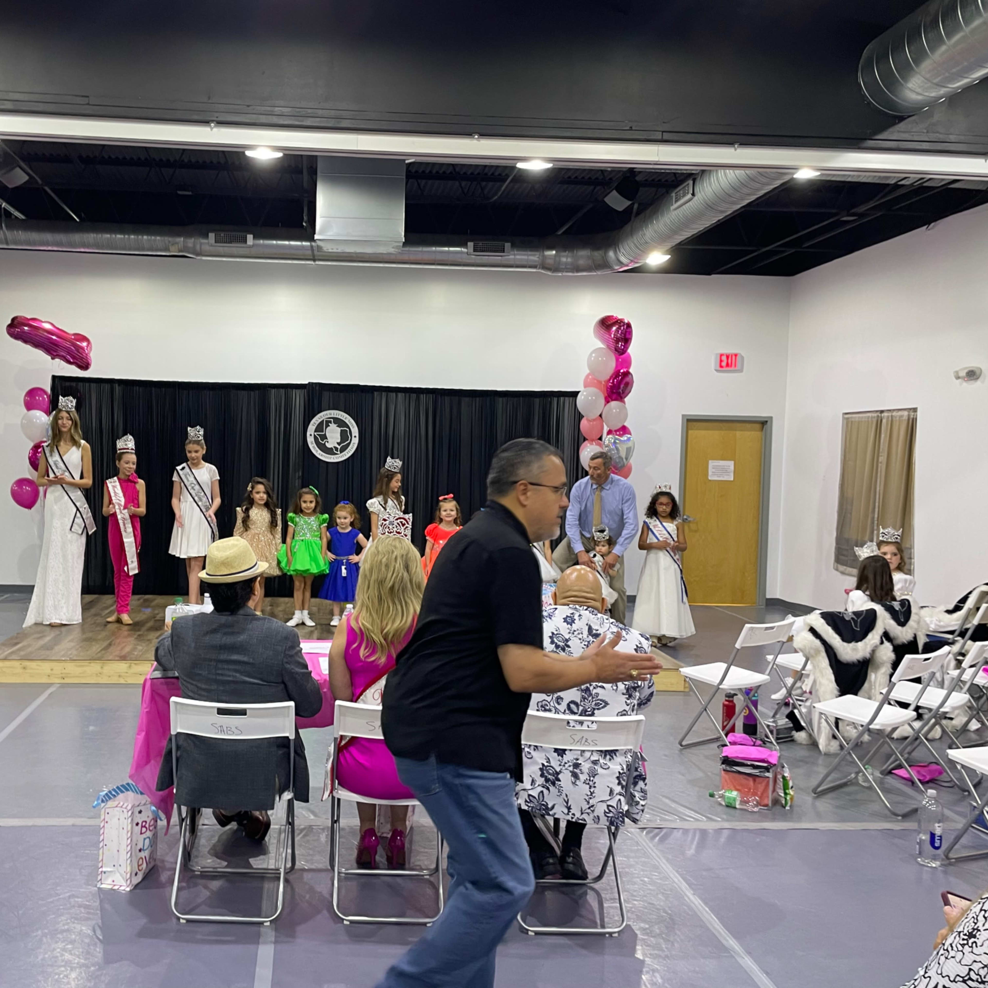 A stage features contestants in formal attire while judges observe from tables, and an audience watches the event in a large room decorated with balloons.