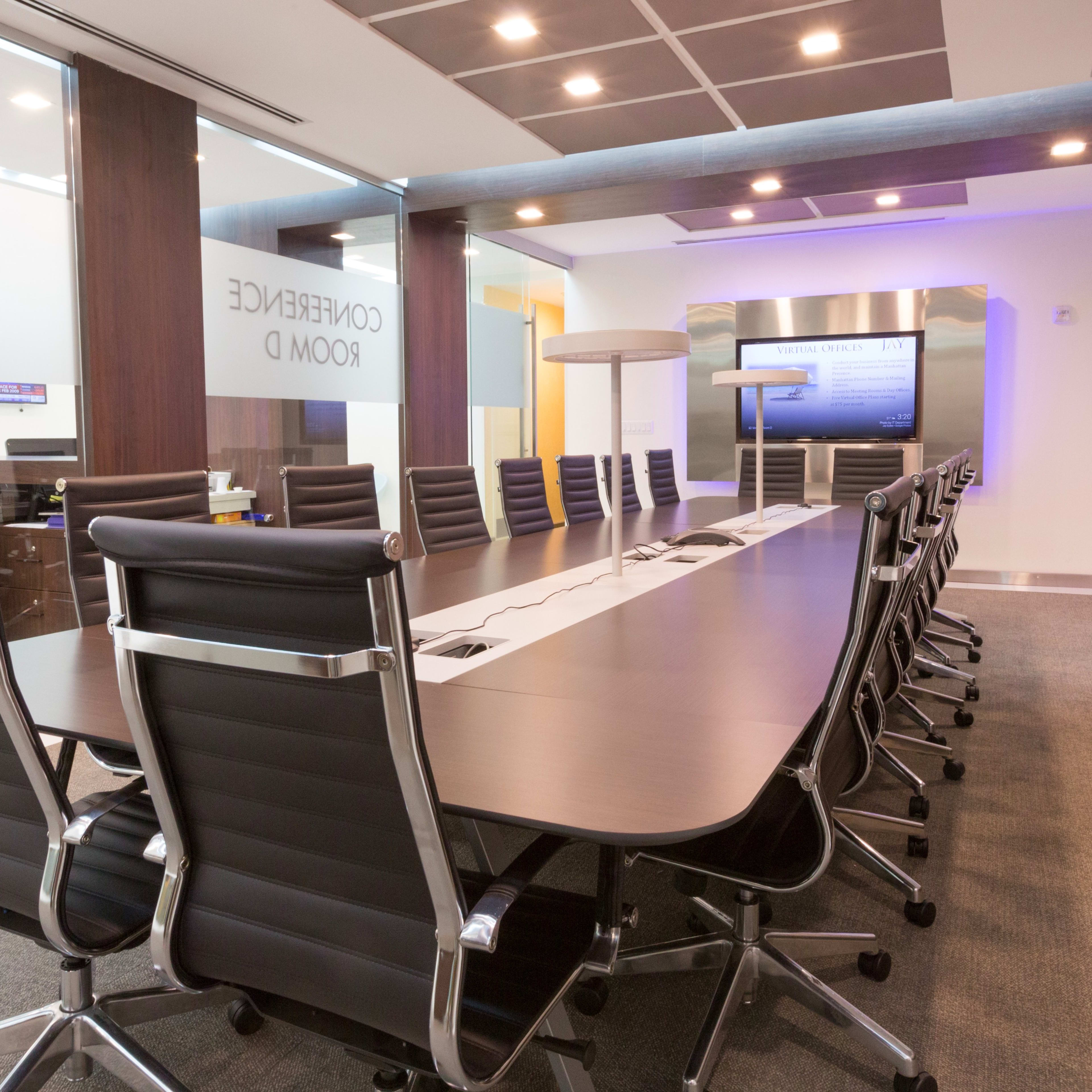 A modern conference room with a long, rectangular table and black ergonomic chairs, complemented by a digital display and decorative flowers.
