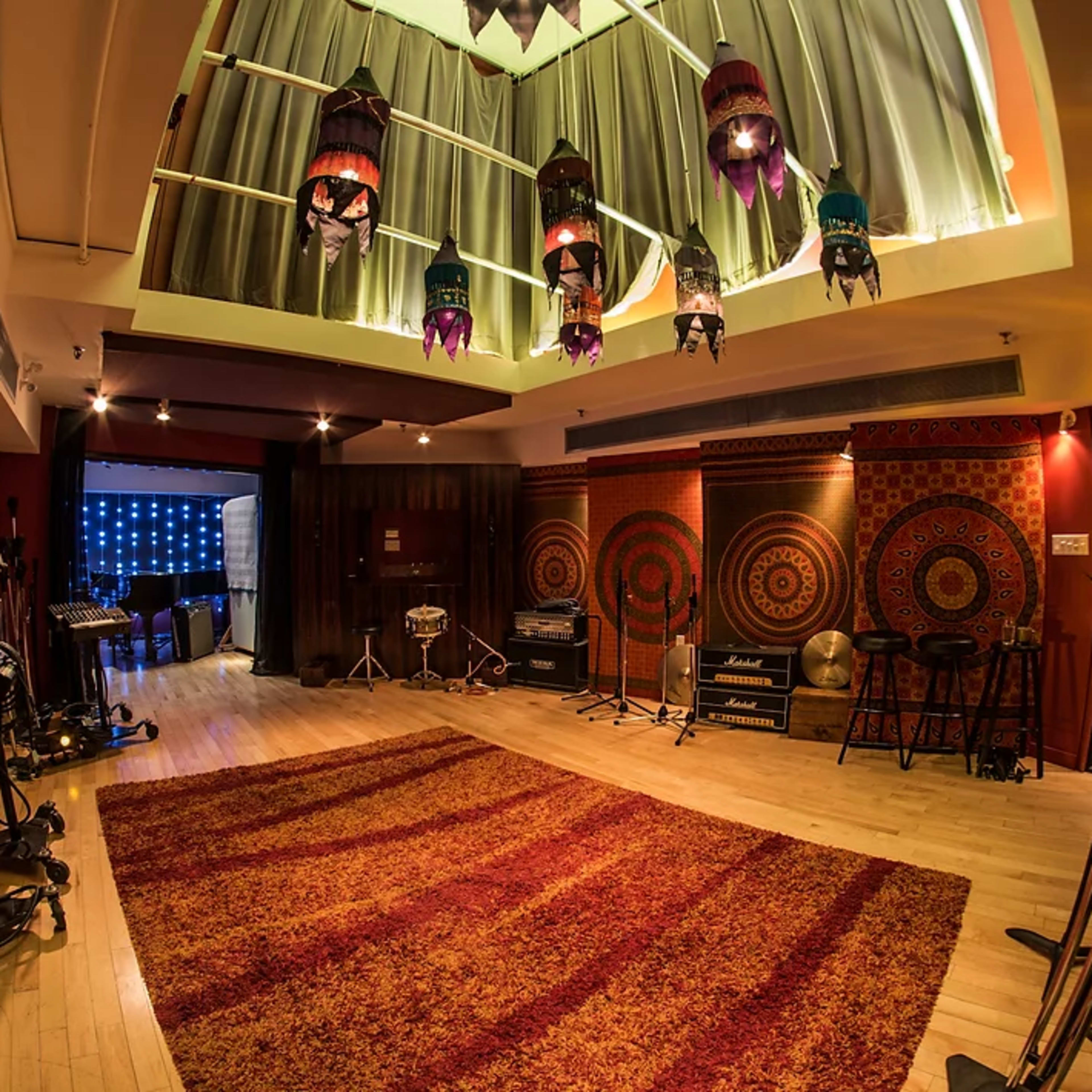 A music recording studio features a spacious room with decorative rugs, microphones, and amplifiers, illuminated by colorful pendant lights.