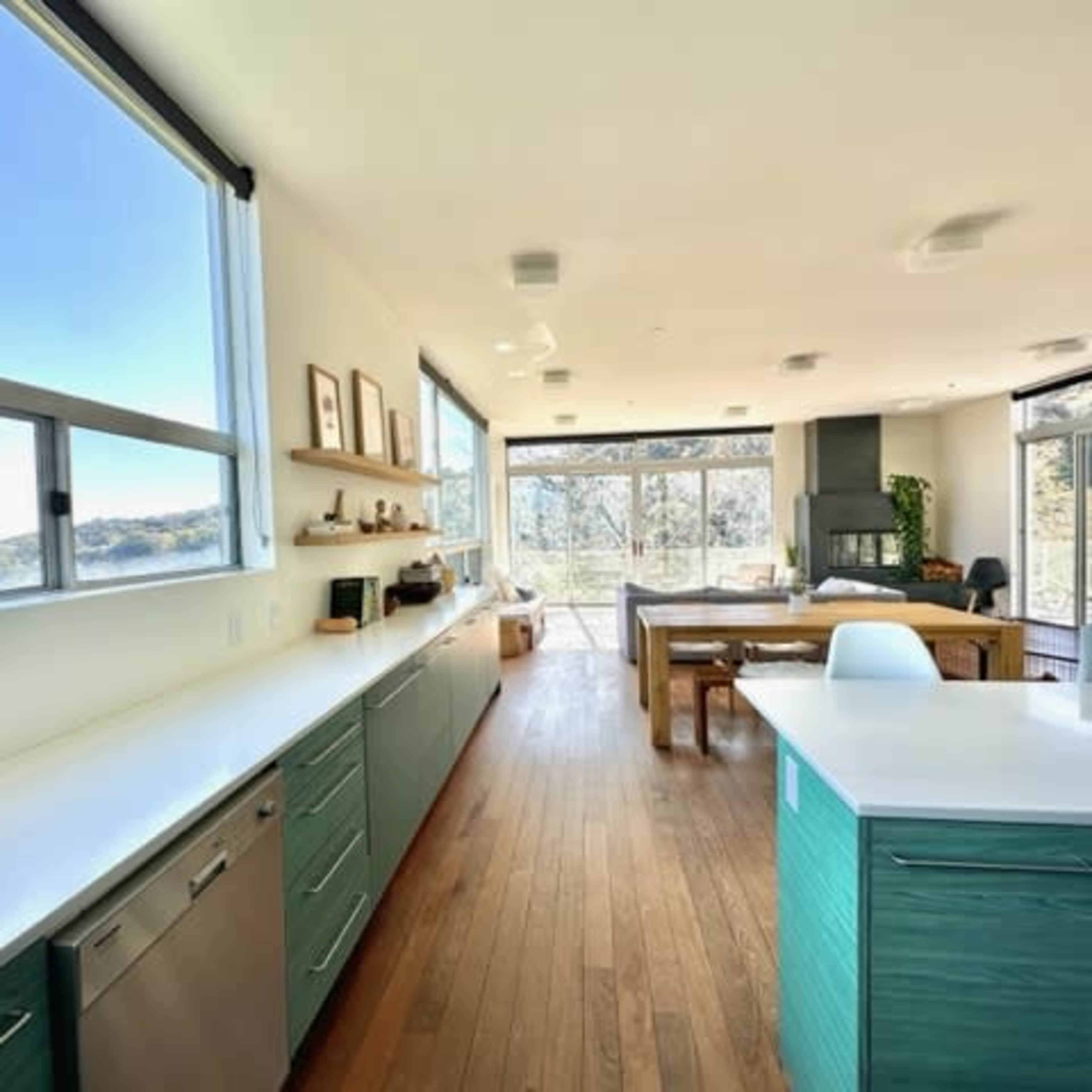 A modern kitchen with green cabinets, a large window providing a view of the outdoors, and an open layout that connects to a dining and living area.