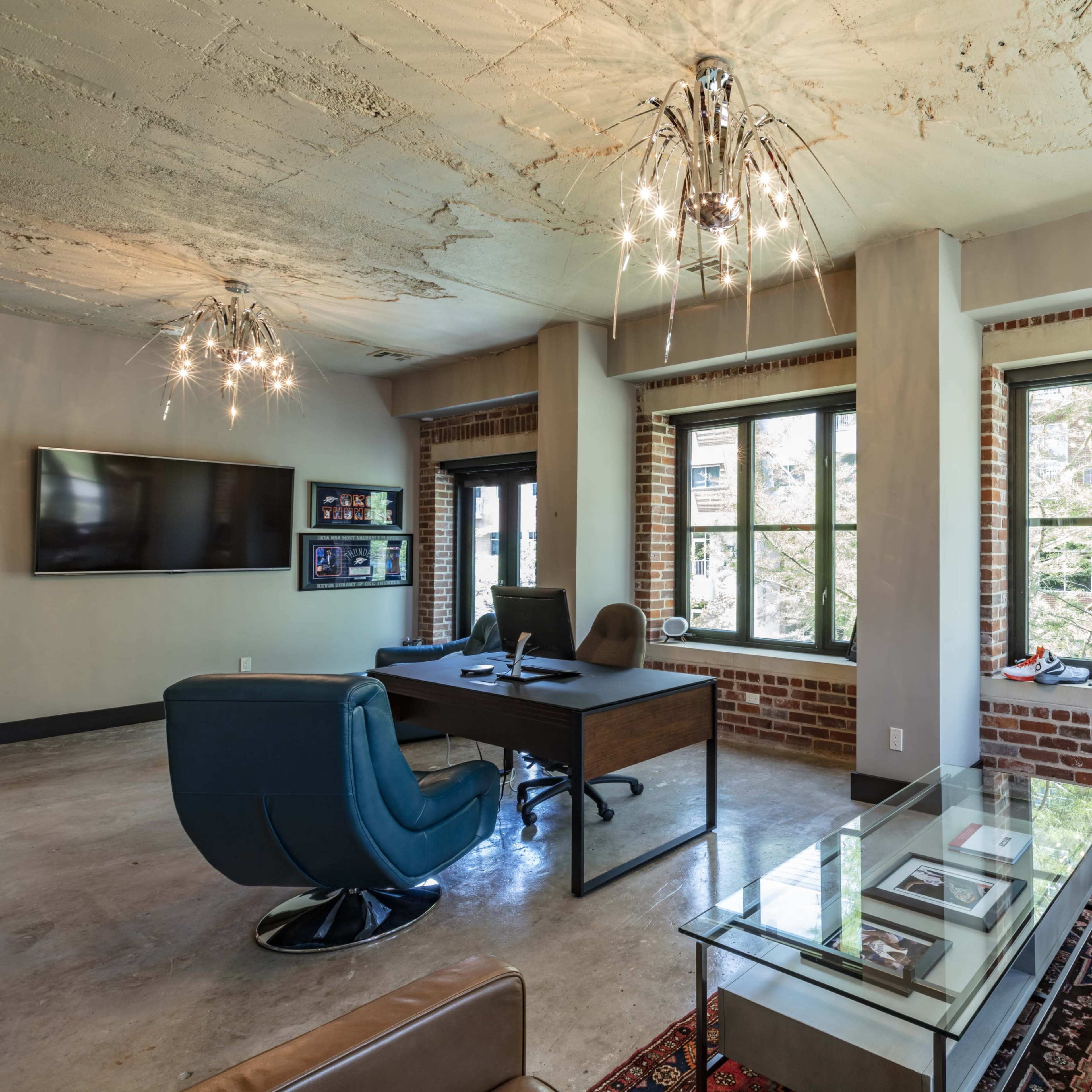 A modern office space with exposed brick walls, a desk and chair, a large television, and a glass coffee table in front of a sofa.