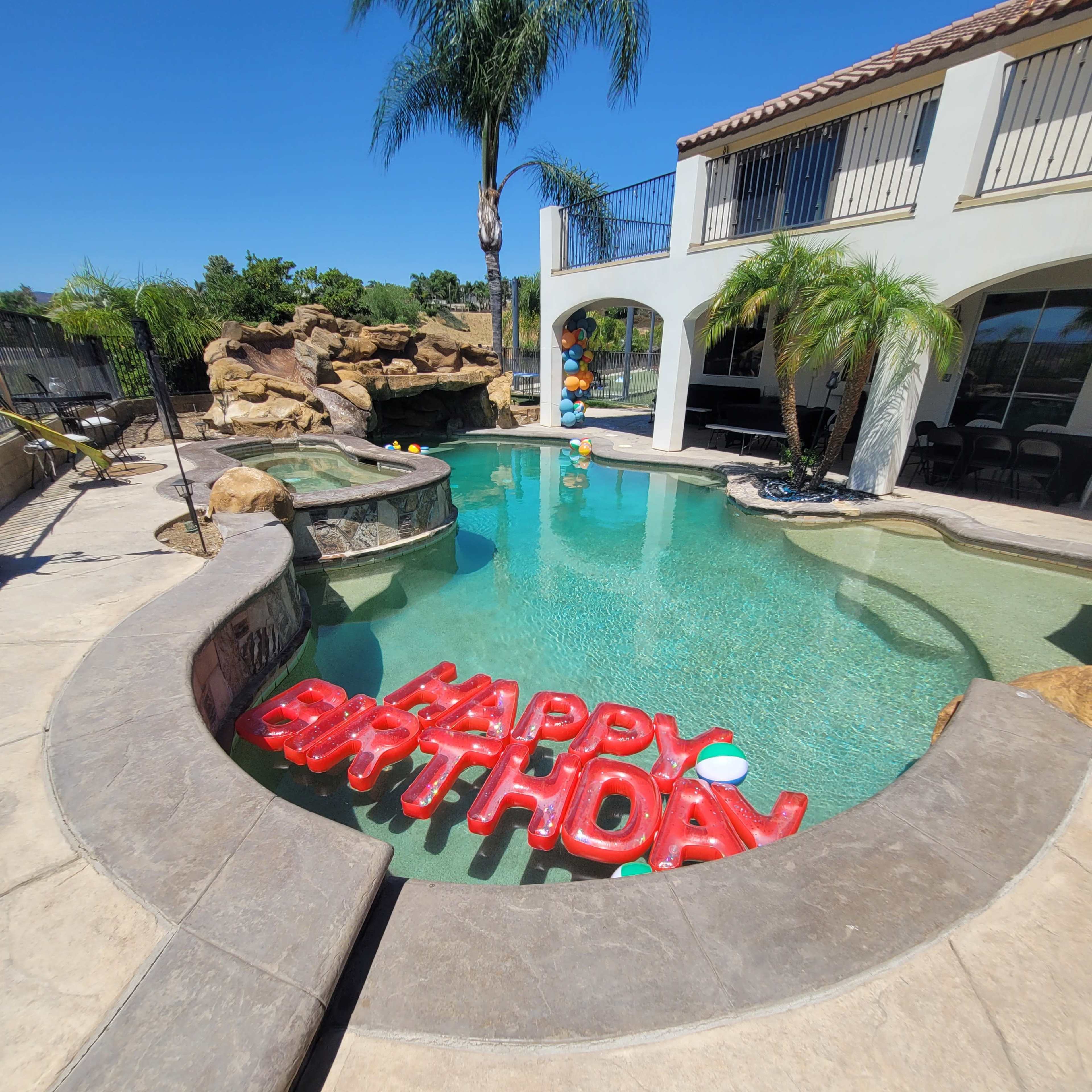 A swimming pool with "HAPPY BIRTHDAY" balloons floats on the water, surrounded by palm trees and a stone waterfall.