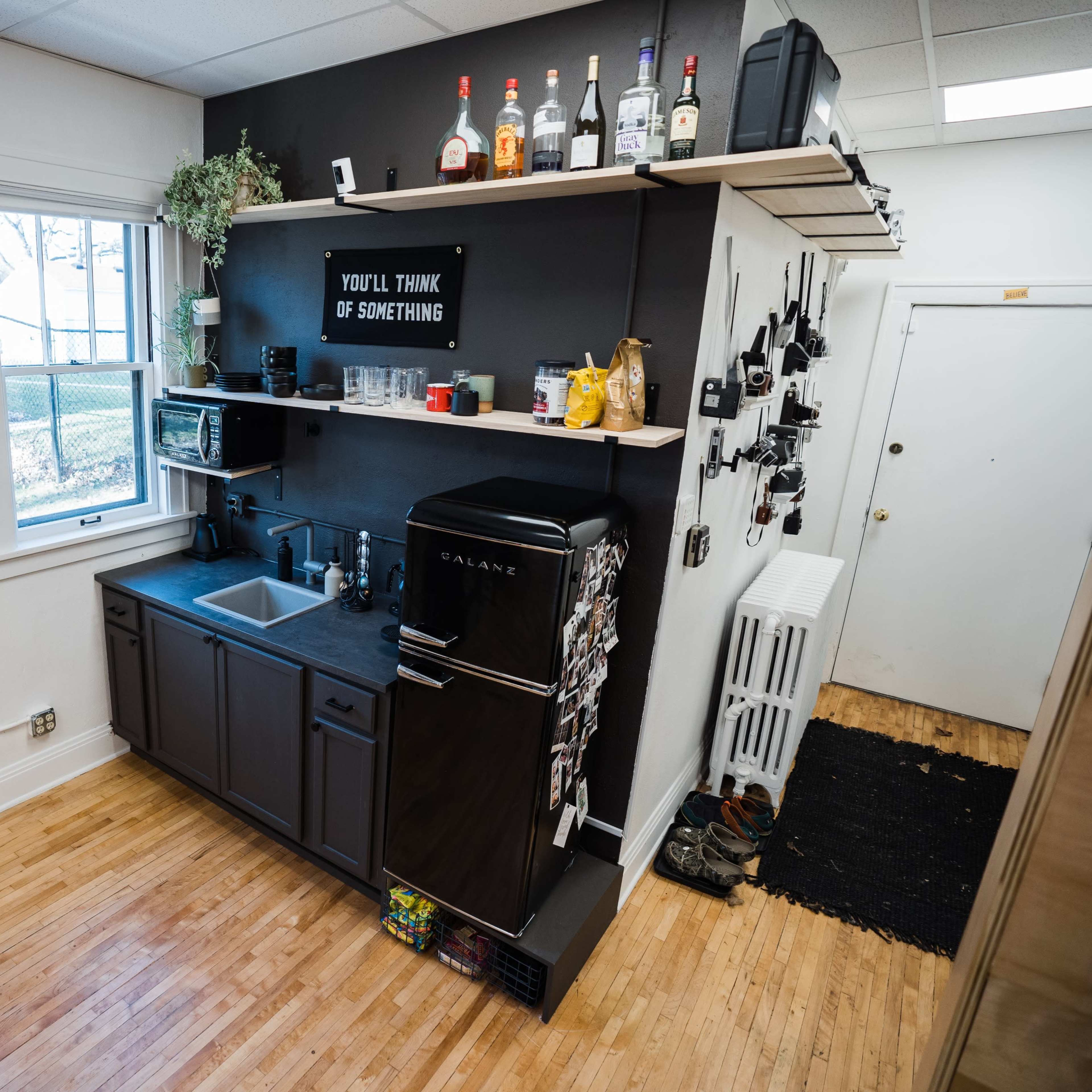 A compact kitchen featuring a black refrigerator, a sink, shelves with various kitchen items, and a window providing natural light.