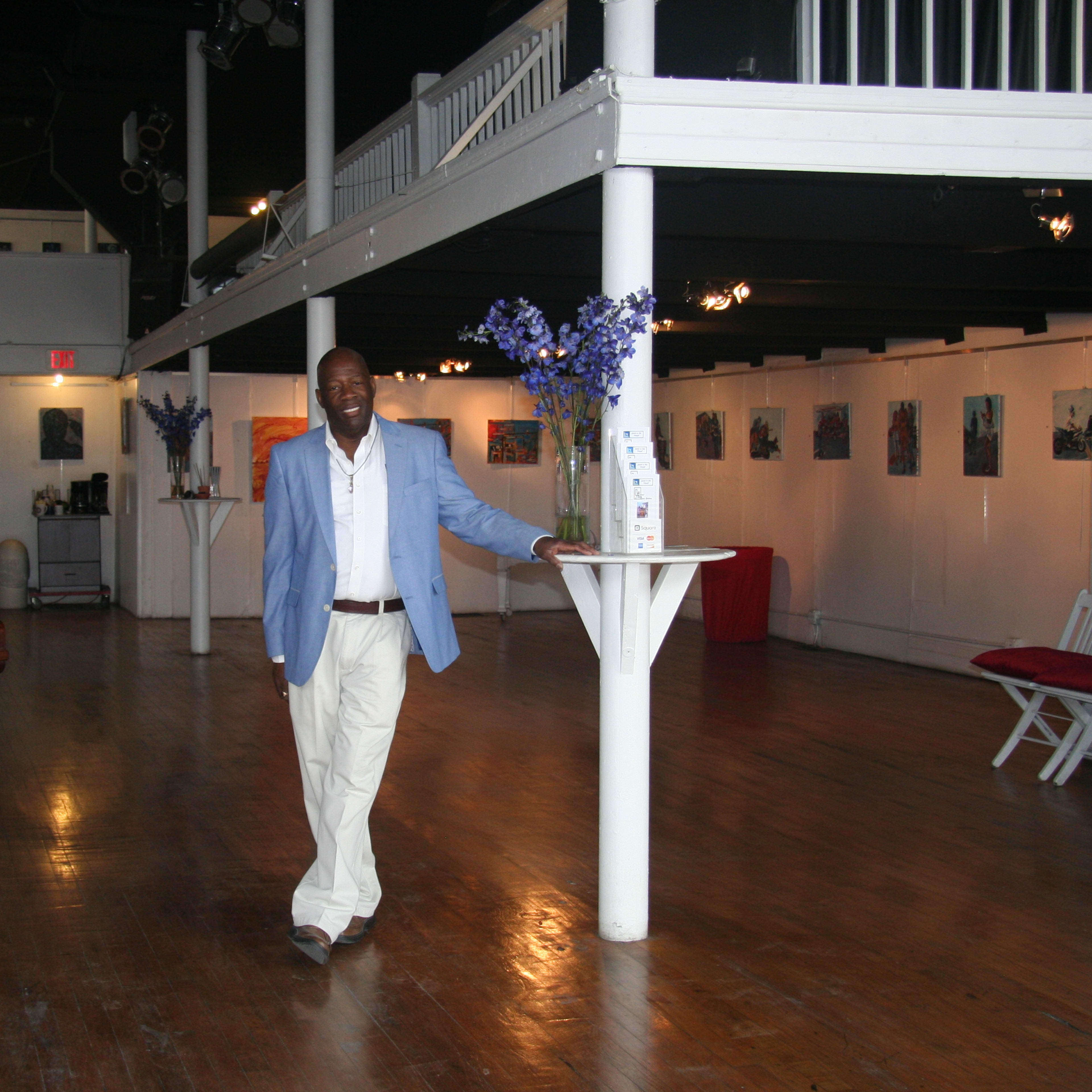 A man in a light blue blazer and white pants stands beside a column in an art gallery with wooden floors and a grand piano.