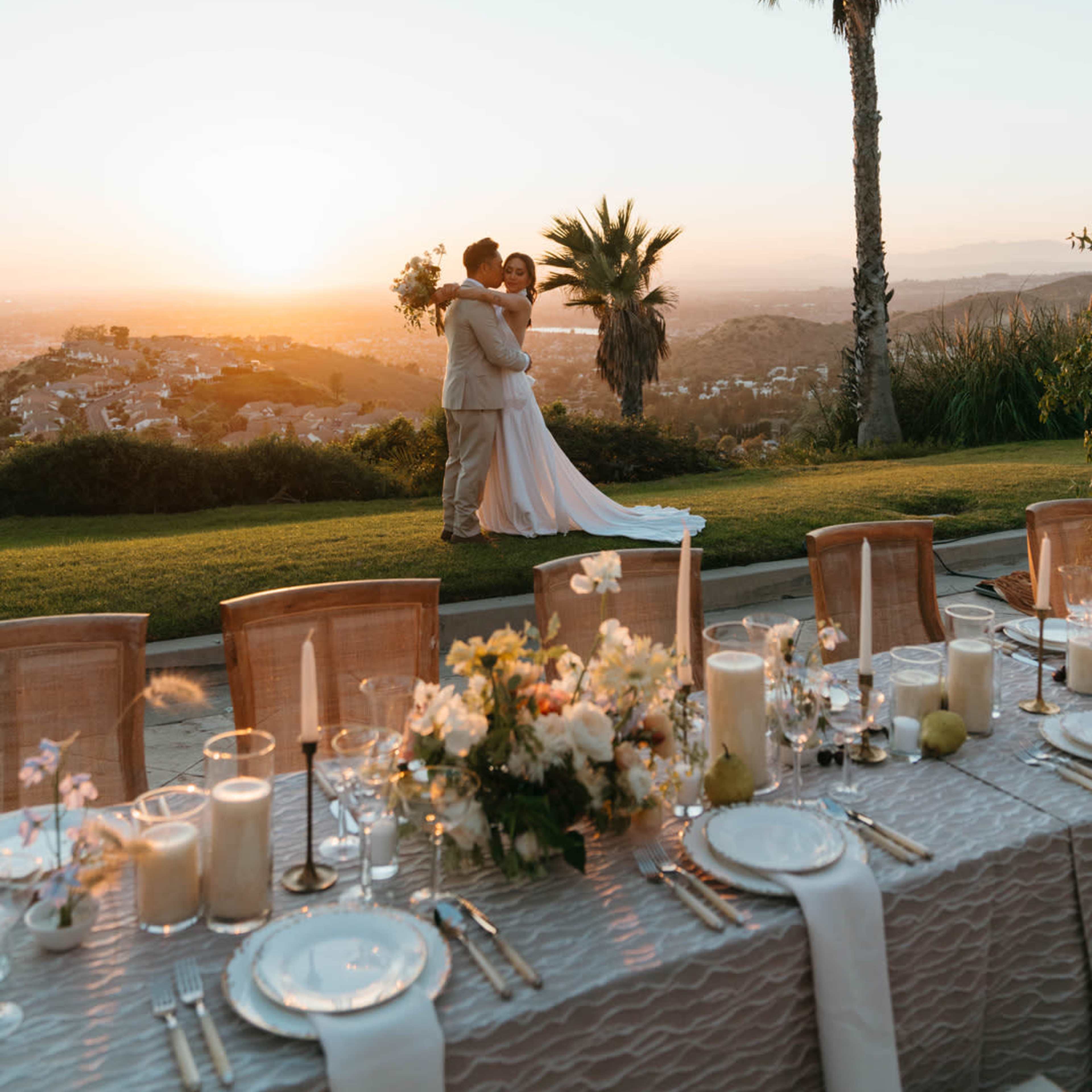 A couple embraces in a wedding setting with a decorated table and a sunset view in the background.