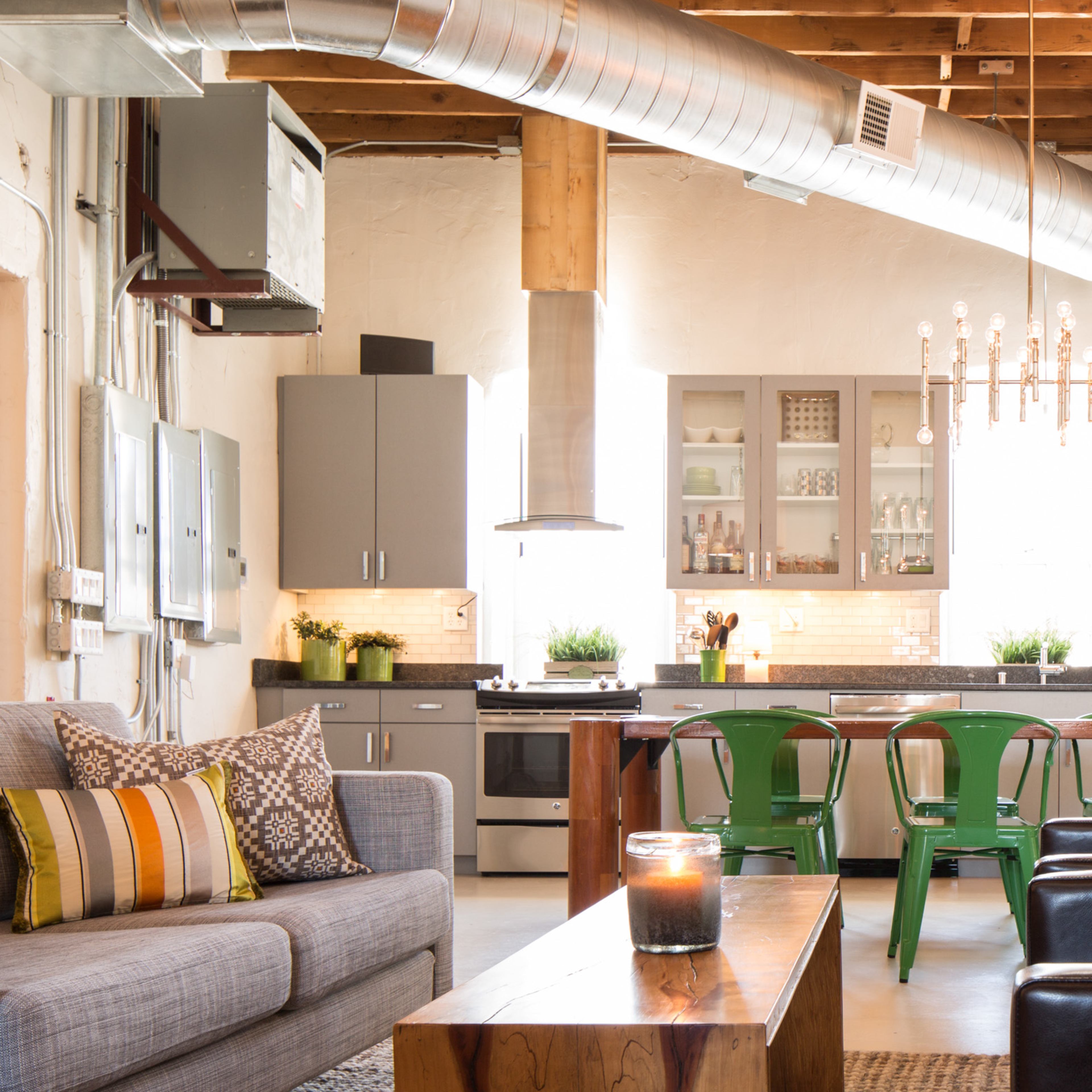 A modern kitchen and living area features stainless steel appliances, green metal chairs, and wooden furniture under exposed beams and ductwork.