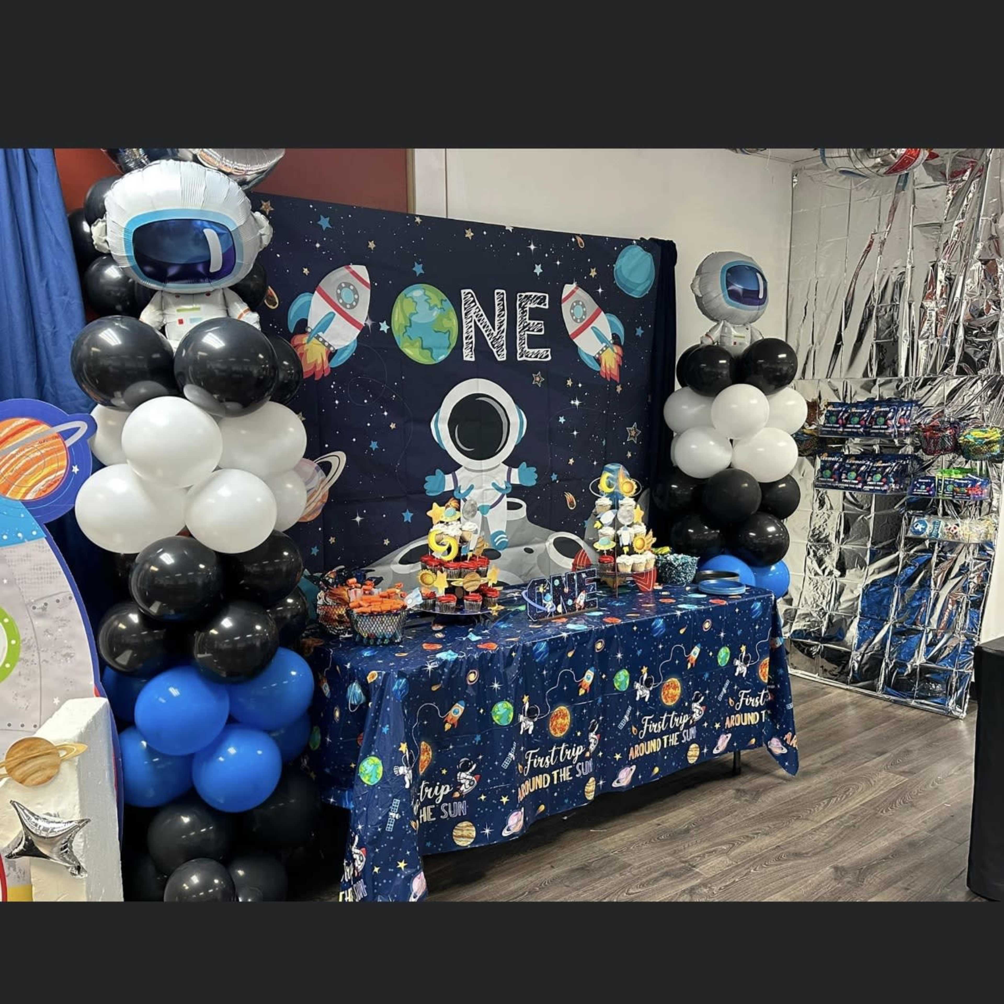 A birthday party setup features a space-themed table adorned with balloons and decorations, displaying a cake and treats under a backdrop that reads "ONE."