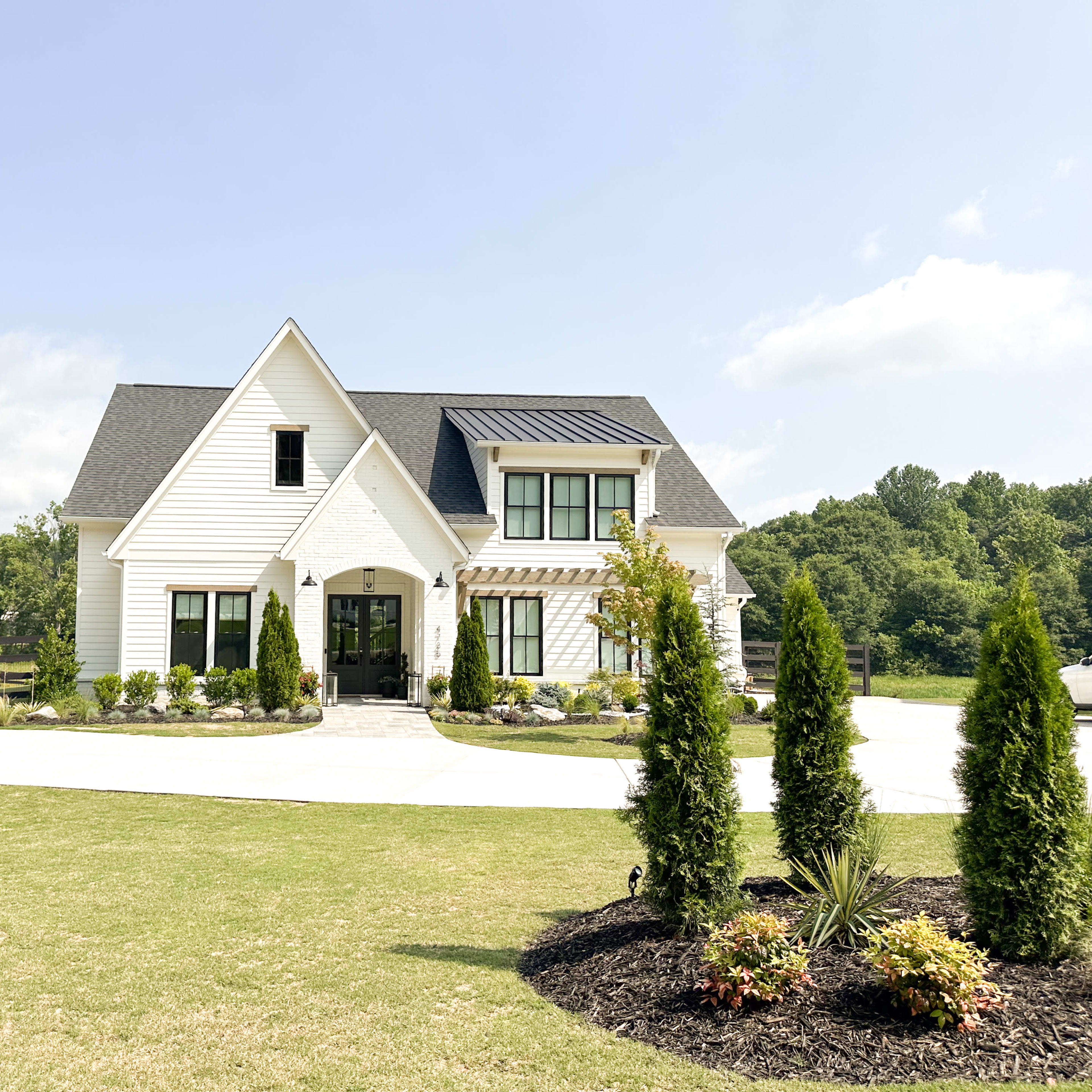 A modern two-story white house with a black roof, surrounded by neatly landscaped green grass and colorful flower beds.