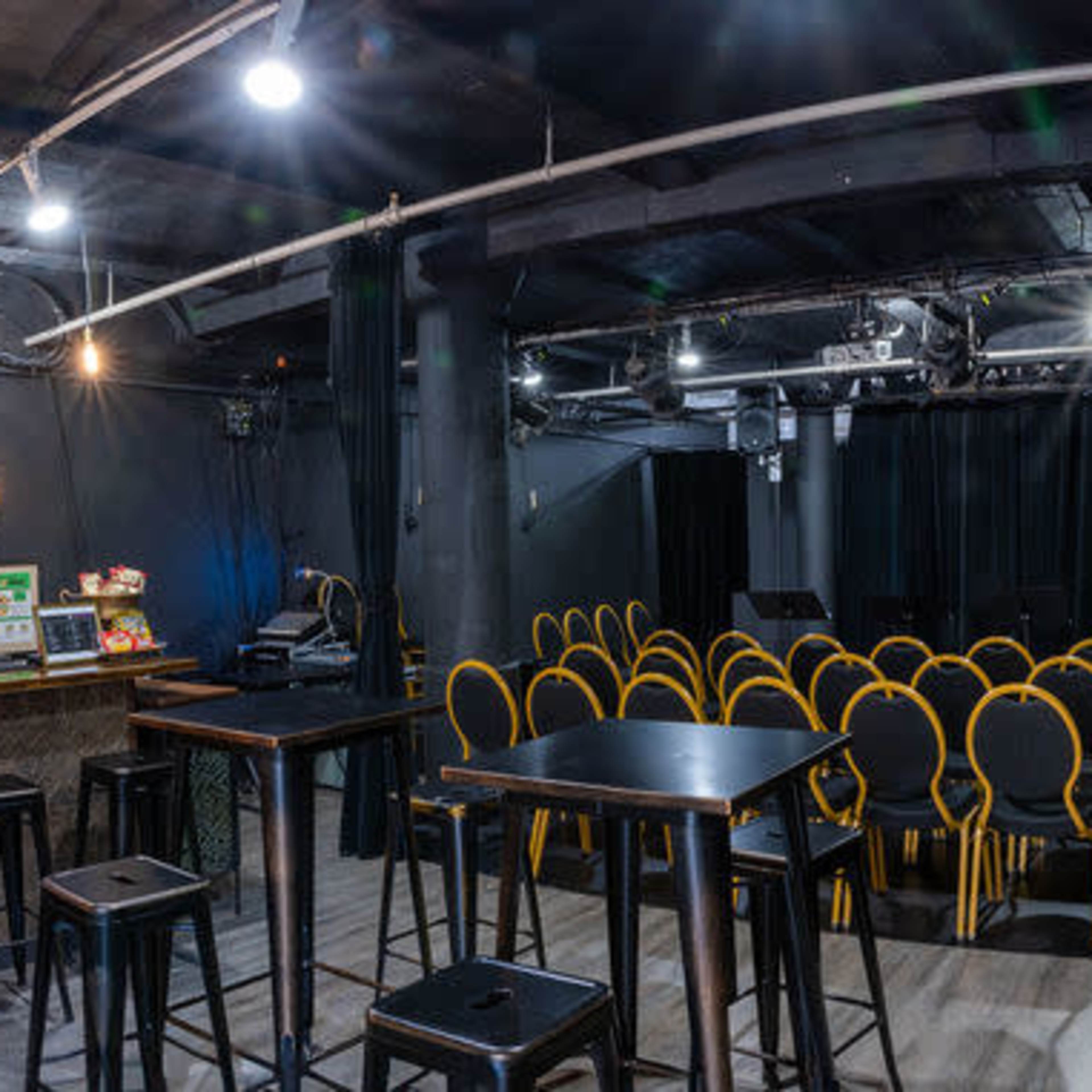 A dimly lit bar area with high tables and stools, a counter with drinks, and a small stage in the background surrounded by rows of yellow chairs.