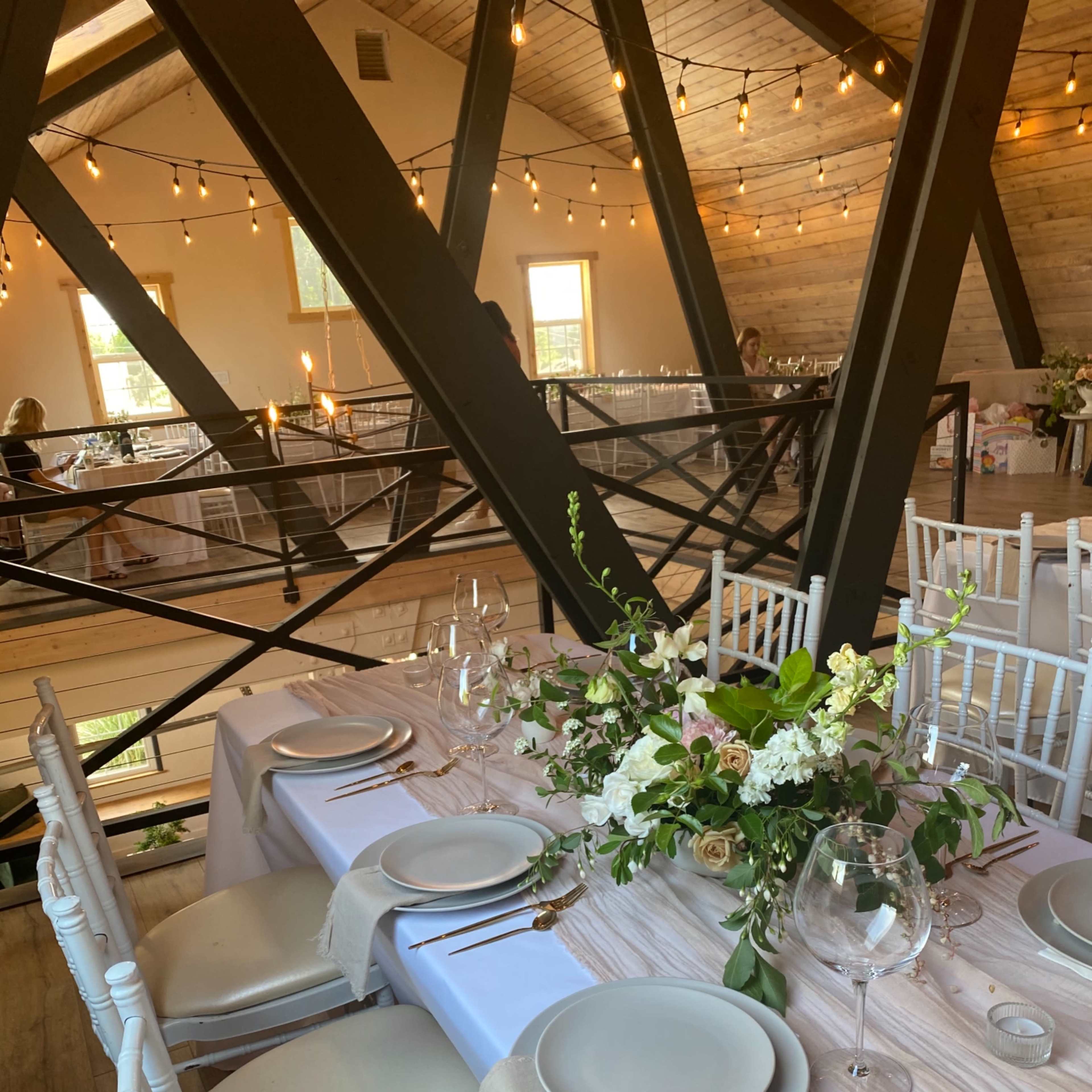 A dining table is elegantly set with plates, glasses, and floral arrangements in a rustic venue adorned with string lights.