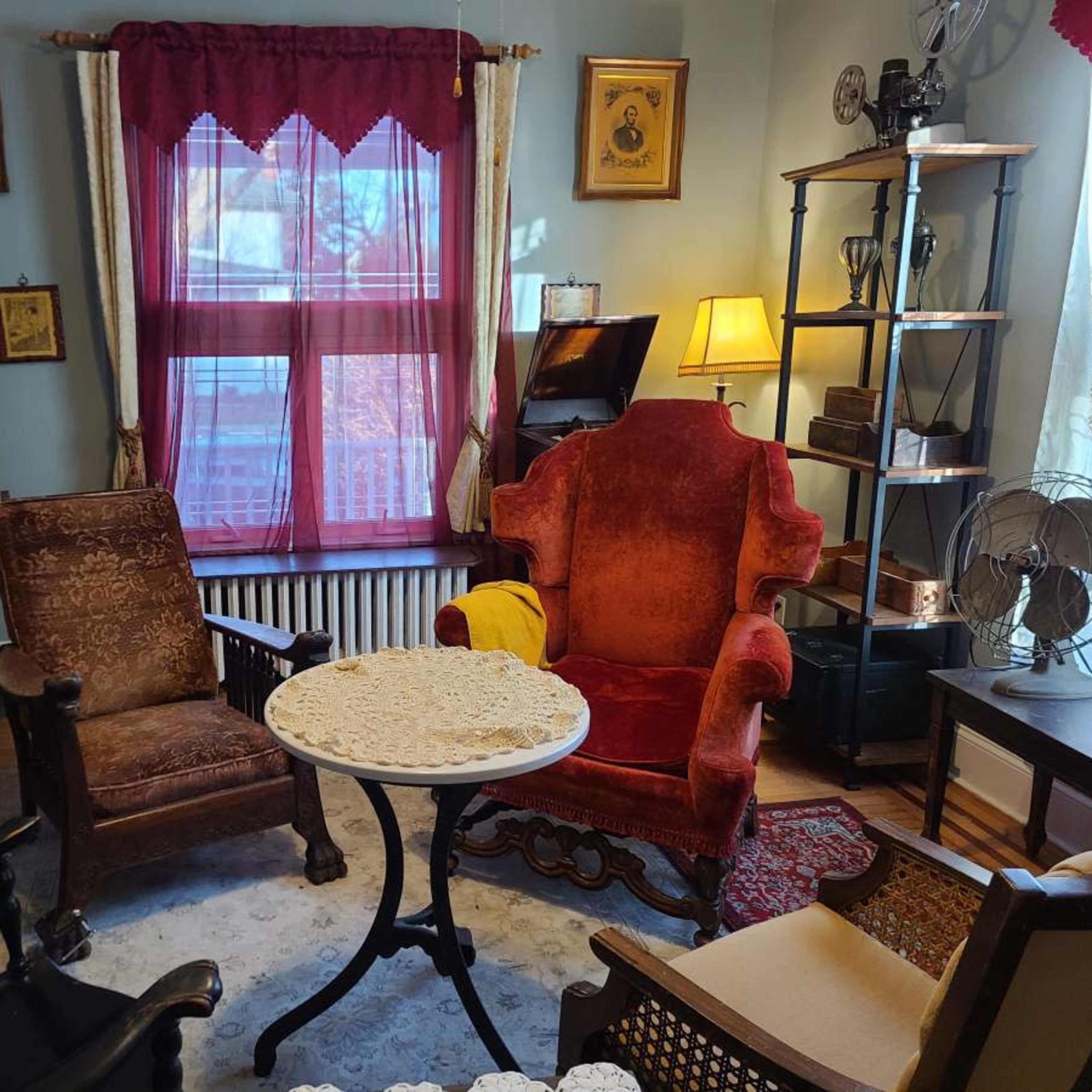 A cozy room furnished with a round table, two upholstered chairs, and various antique decorations, illuminated by natural light coming through a window.