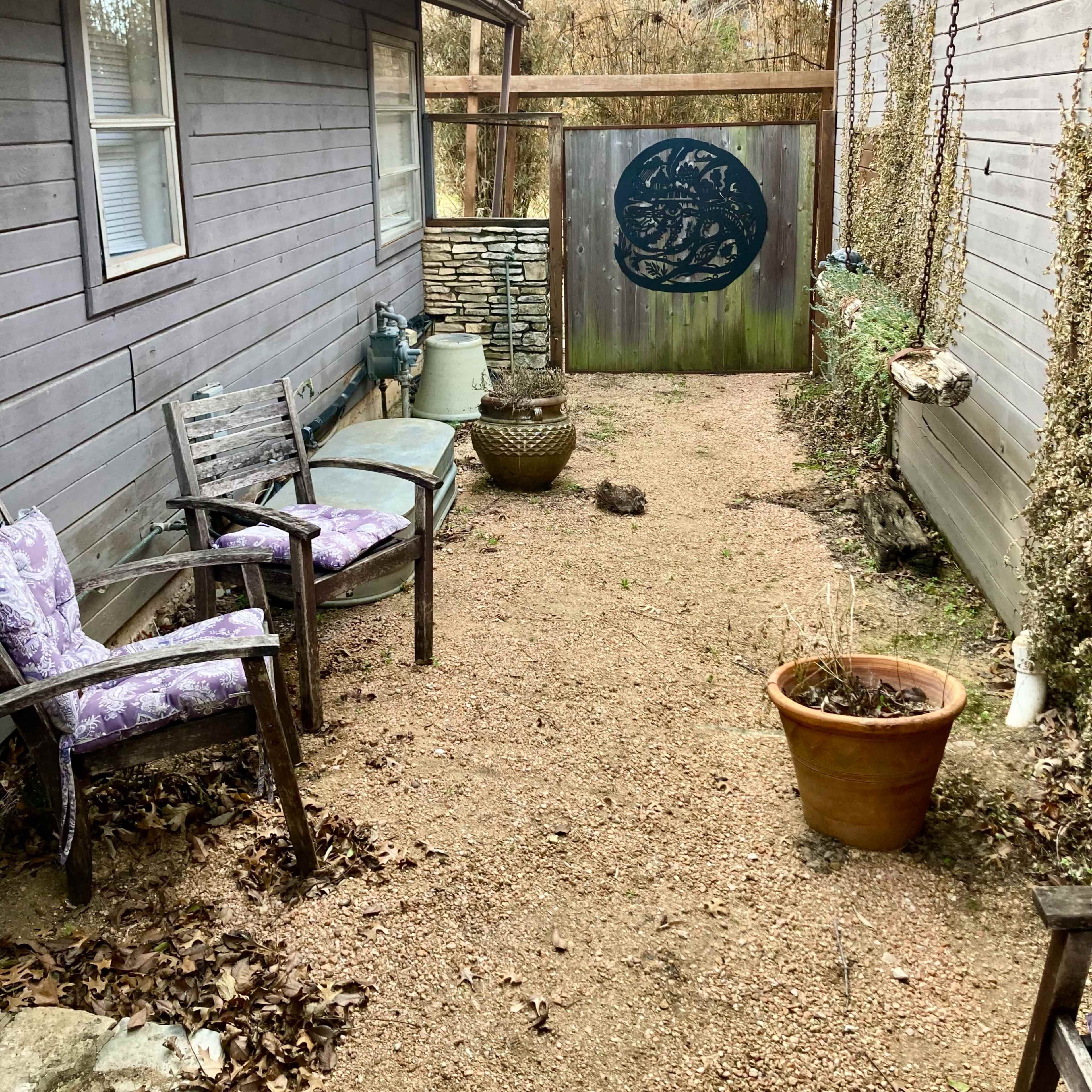 A narrow pathway lined with gravel runs between two wooden buildings, featuring a decorative gate at the far end and a couple of chairs on one side.