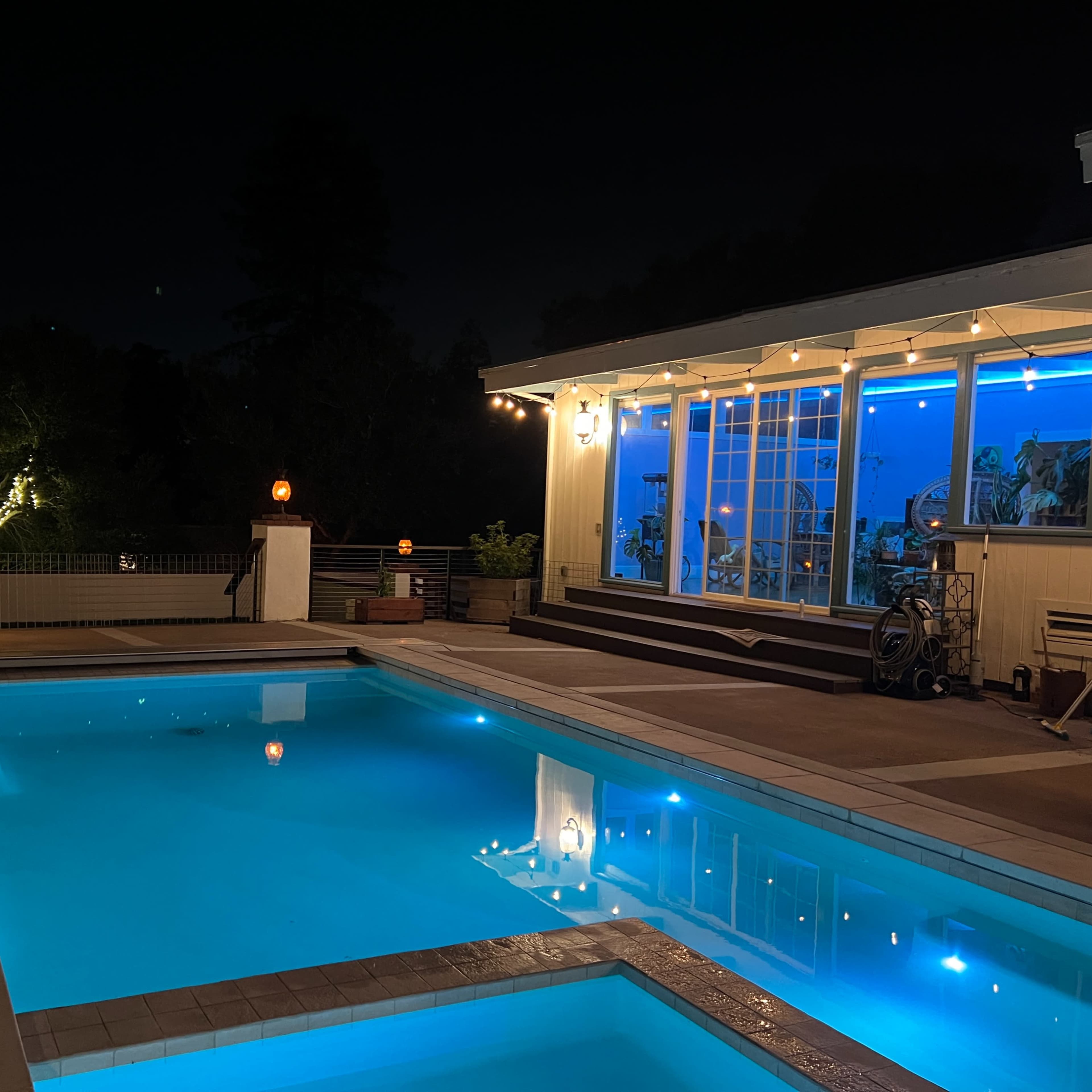 A well-lit swimming pool is surrounded by a wooden deck and a house with large windows, set against a night sky.