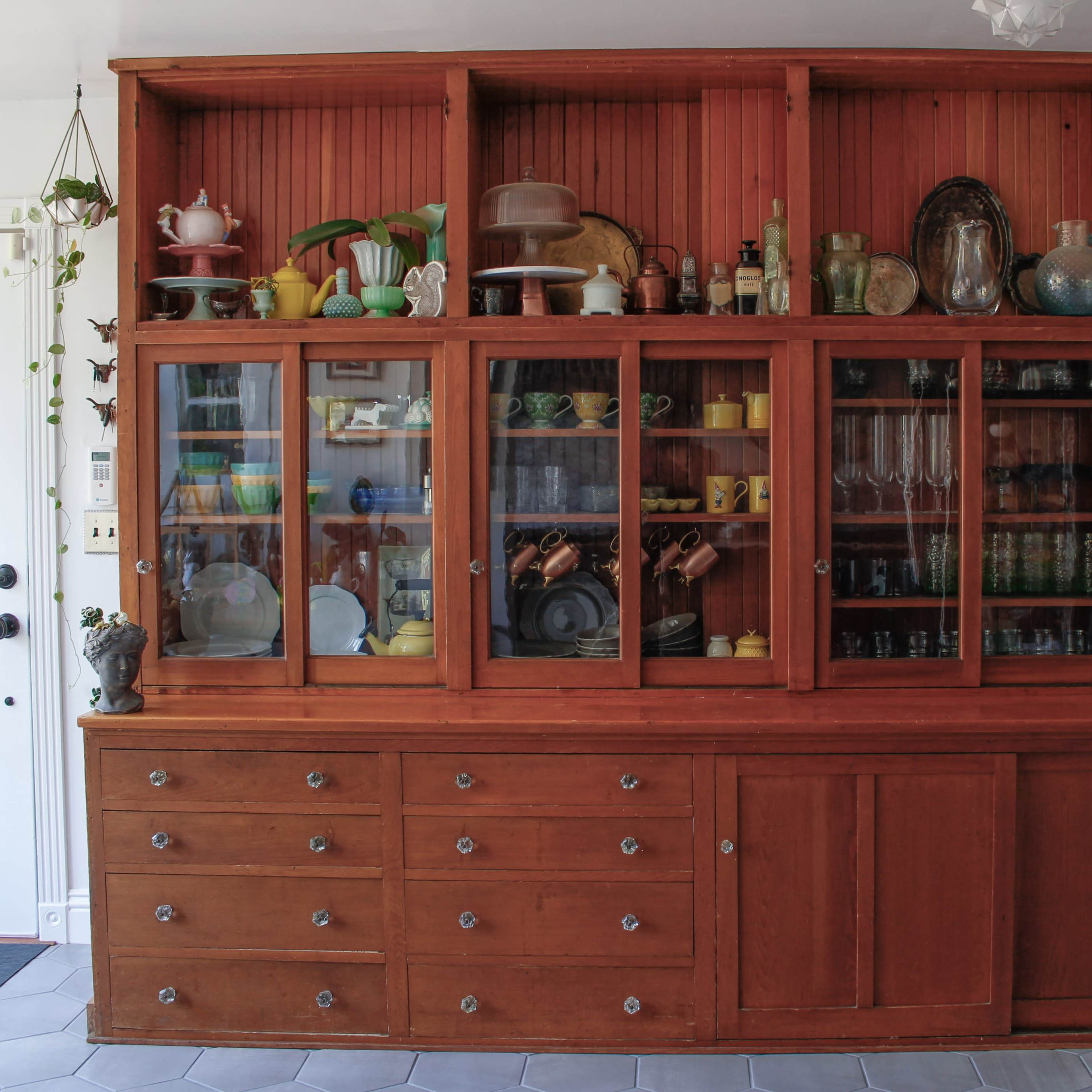 A wooden cabinet with glass doors displaying various dishes, glasses, and kitchen items, set against a white wall near a doorway.
