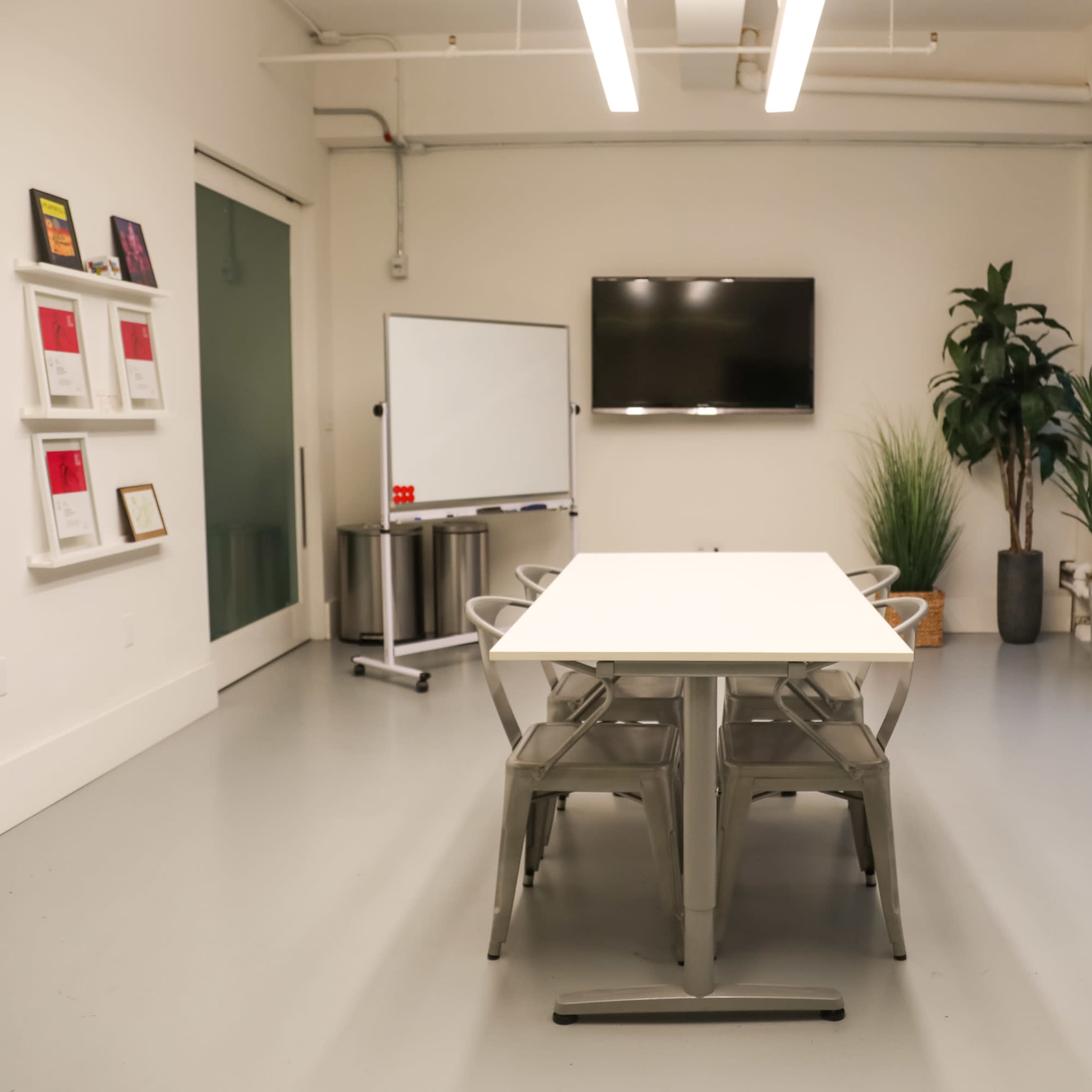 A modern conference room features a long table surrounded by metal chairs, a television mounted on the wall, a whiteboard, and large windows with plants nearby.
