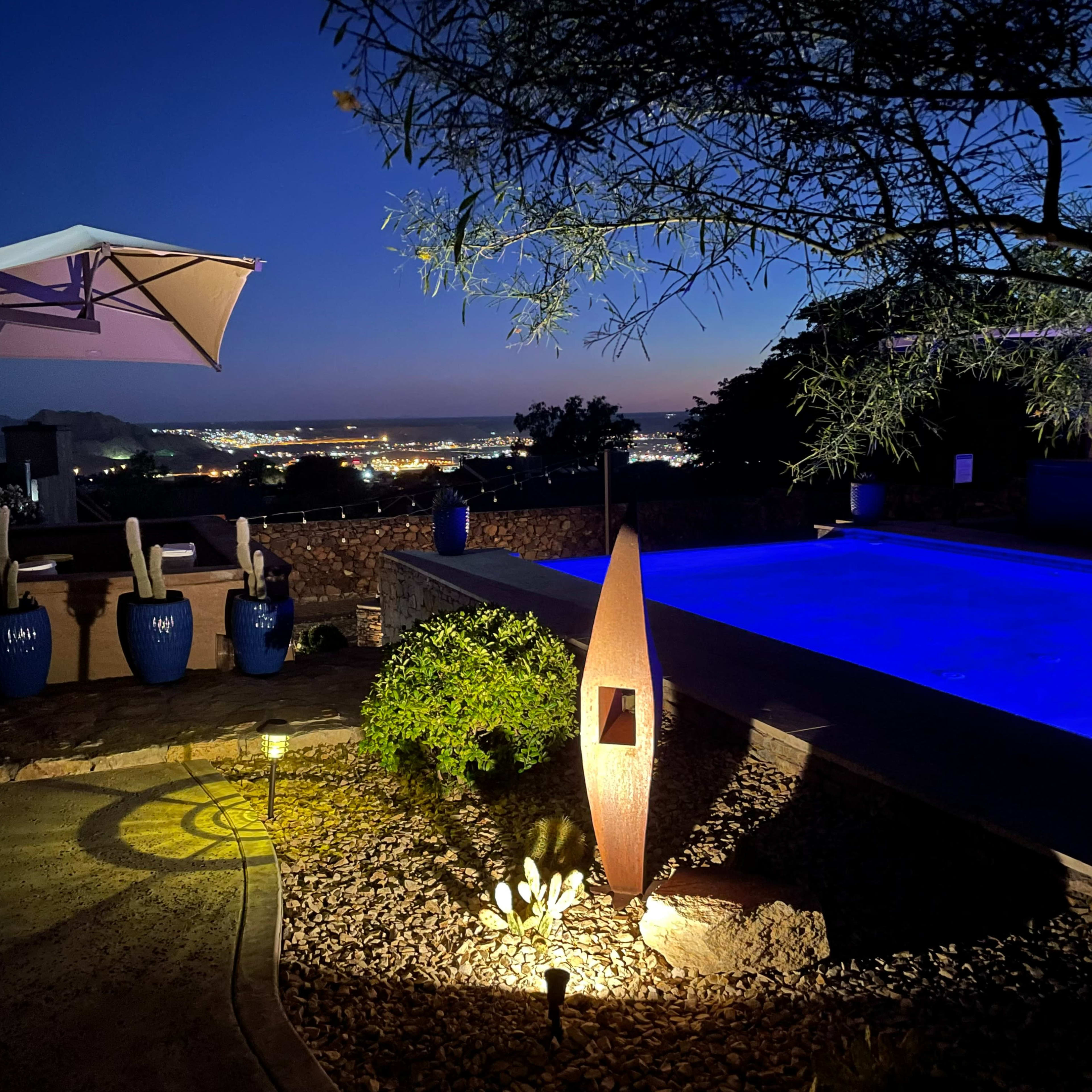 The scene depicts a poolside area at dusk with a modern sculpture, landscaped stones, and city lights visible in the background.